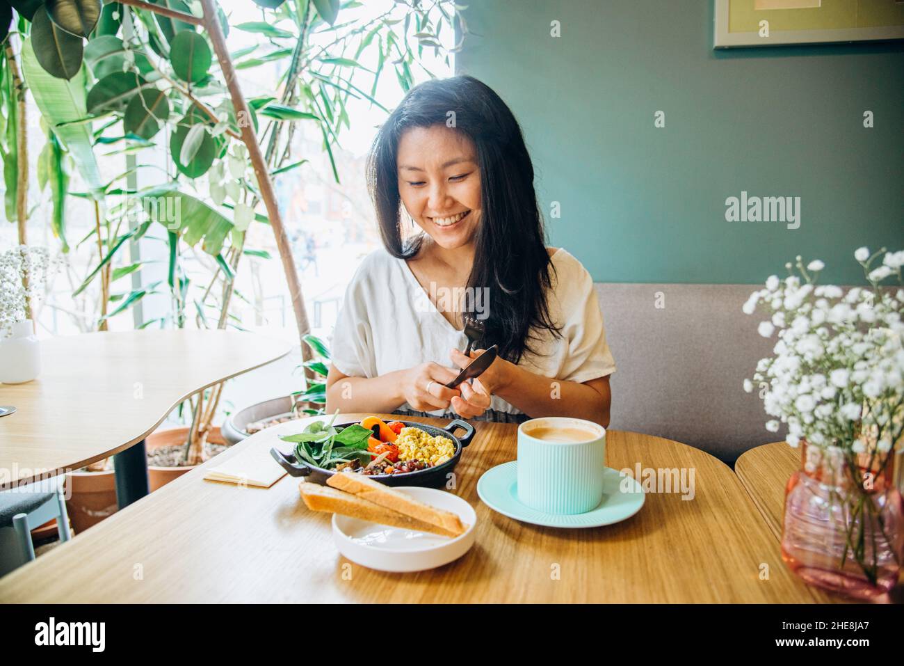 Young Asian woman with long loose black hair sits on sofa at wooden table and eats vegetable dish smiling in cosy vegan cafe closeup Stock Photo