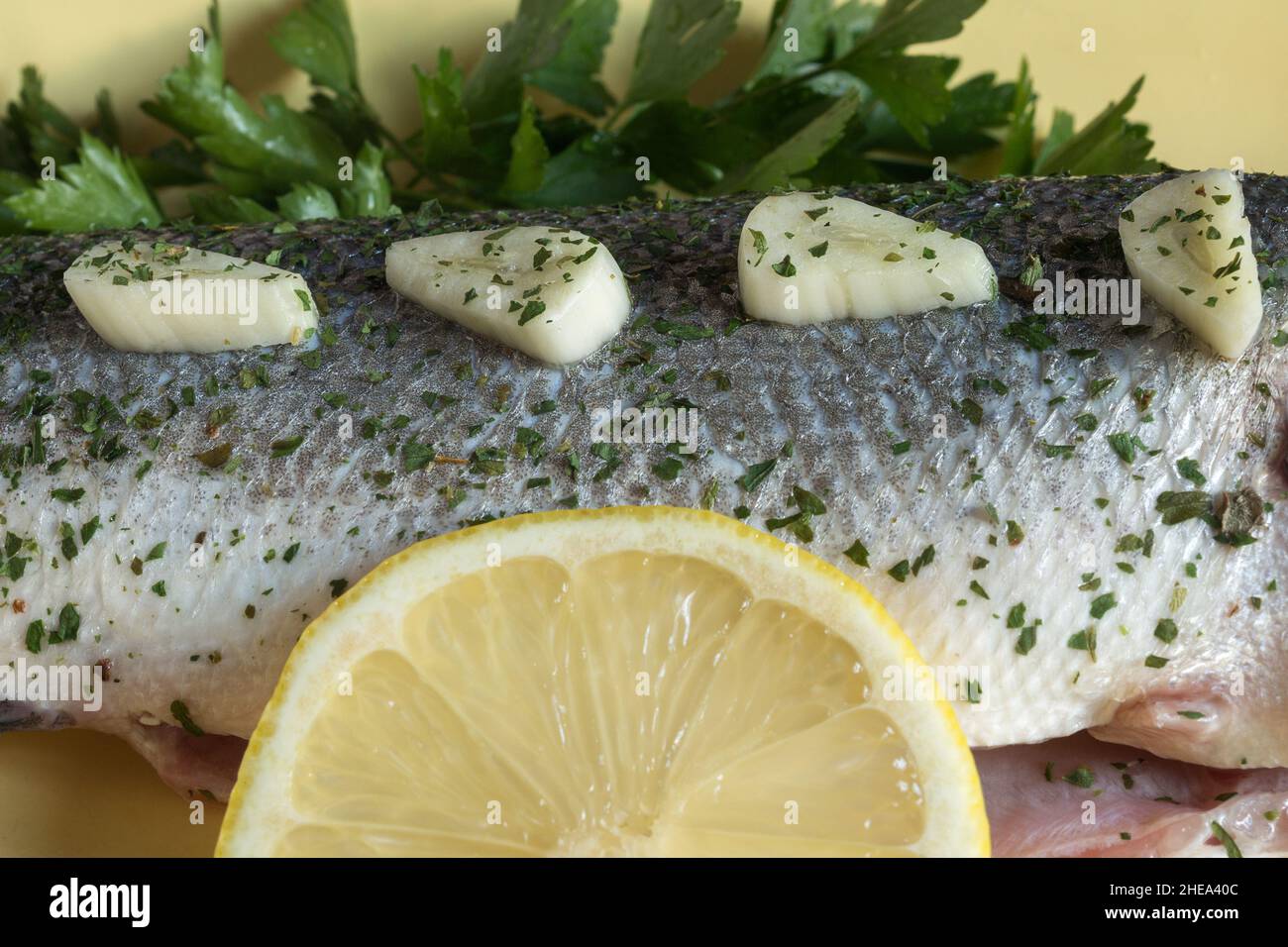 Chunks of garlic seasoned with spice in a row on the skin of raw sea bass accompanied by sprigs of fresh parsley and a lemon slice. Preparation fish. Stock Photo