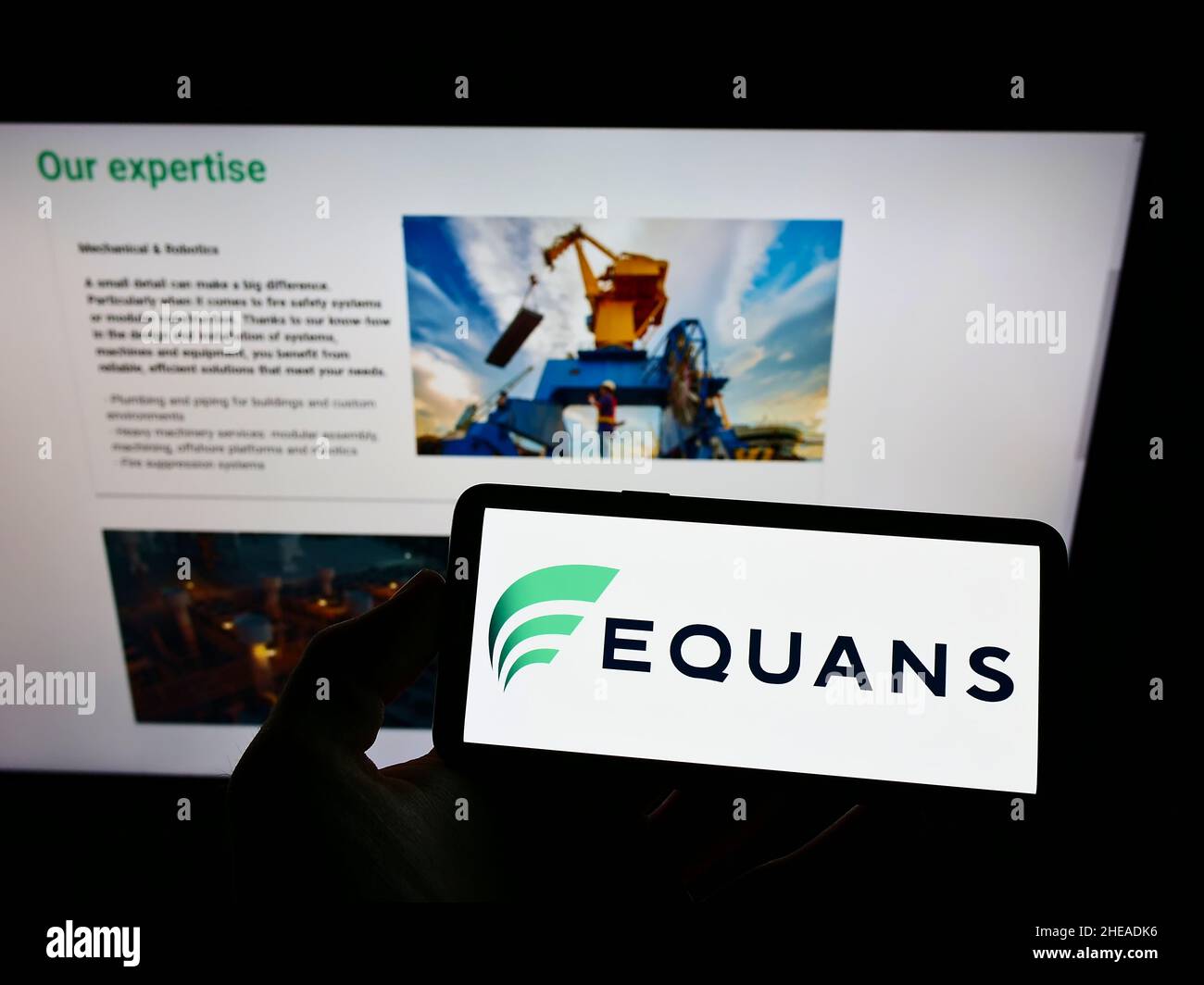 Person holding mobile phone with logo of French facility management company EQUANS on screen in front of business web page. Focus on phone display. Stock Photo