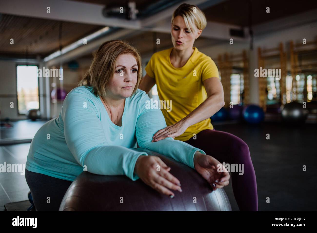 Happy overweight woman exercising with personal trainer on fintess ball in gym Stock Photo