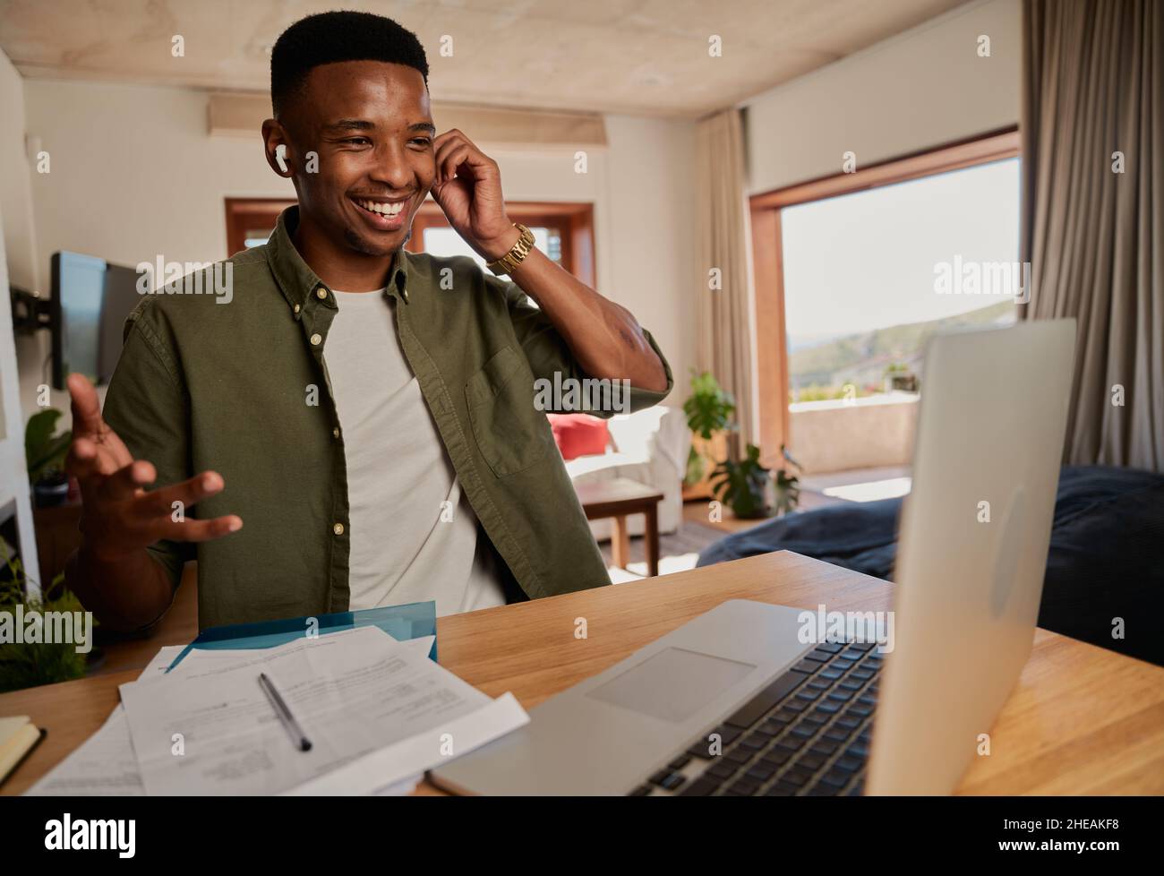 Young adult black male adjusting earphones while talking in online meeting. Sitting at kitchen counter using laptop. Stock Photo