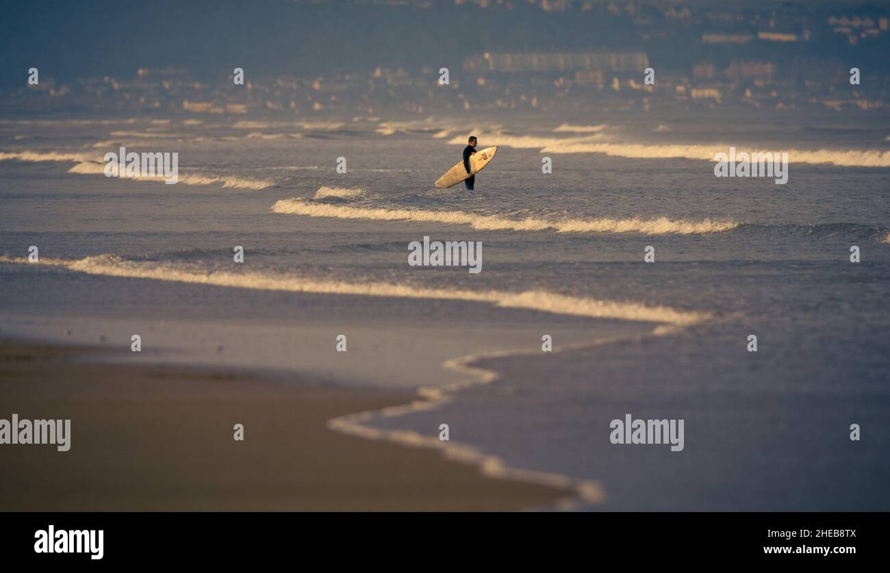 A single surfer with board under arm waits stood in the surf watching the waves on Saunton Sands Beach north Devon. Stock Photo