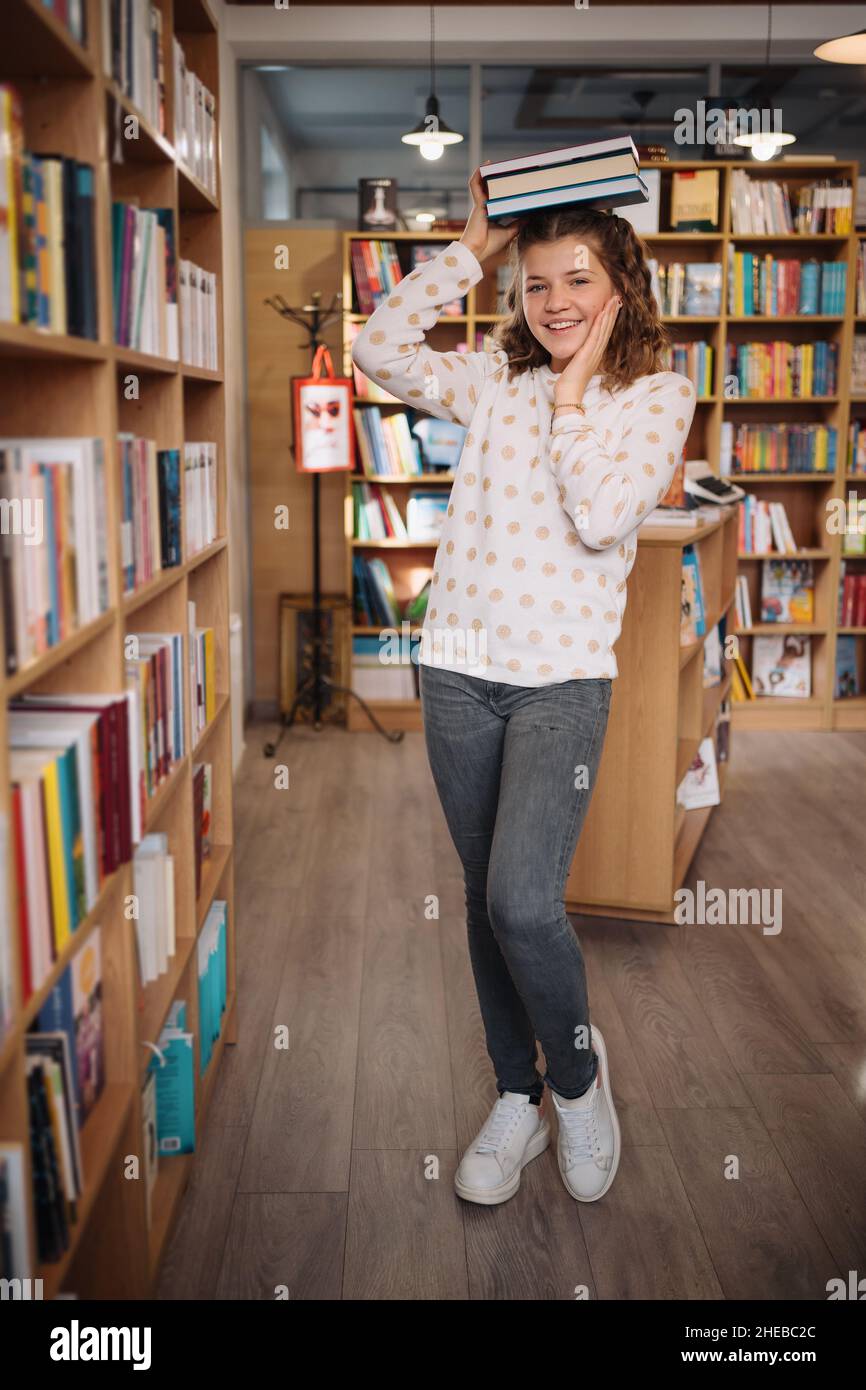 Teen girl among a pile of books. A young girl holding books on head with shelves in the background. She is surrounded by stacks of books. Book day. Stock Photo