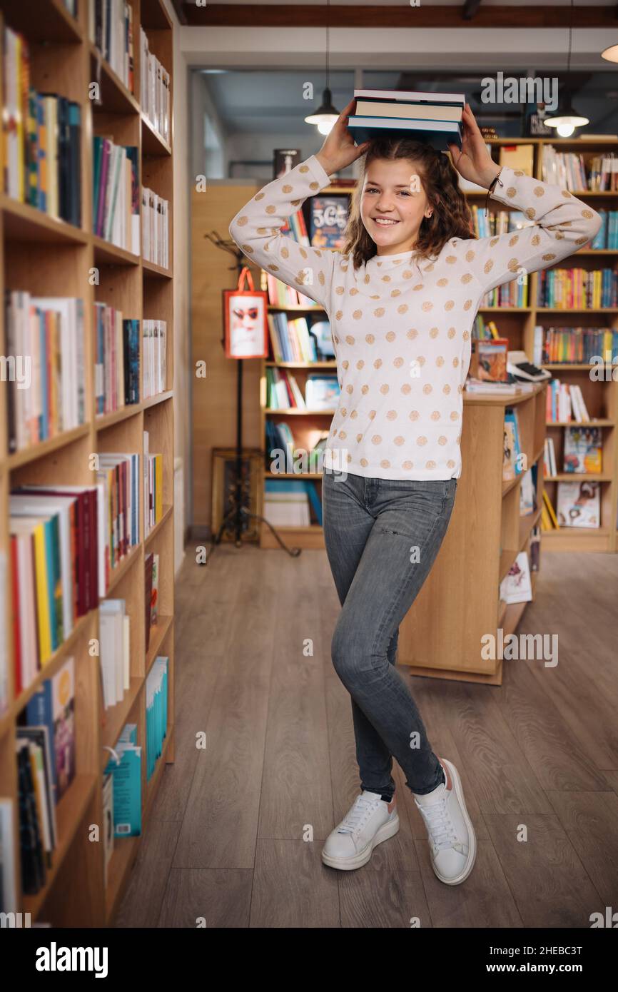 Teen girl among a pile of books. A young girl holding books on head with shelves in the background. She is surrounded by stacks of books. Book day. Stock Photo