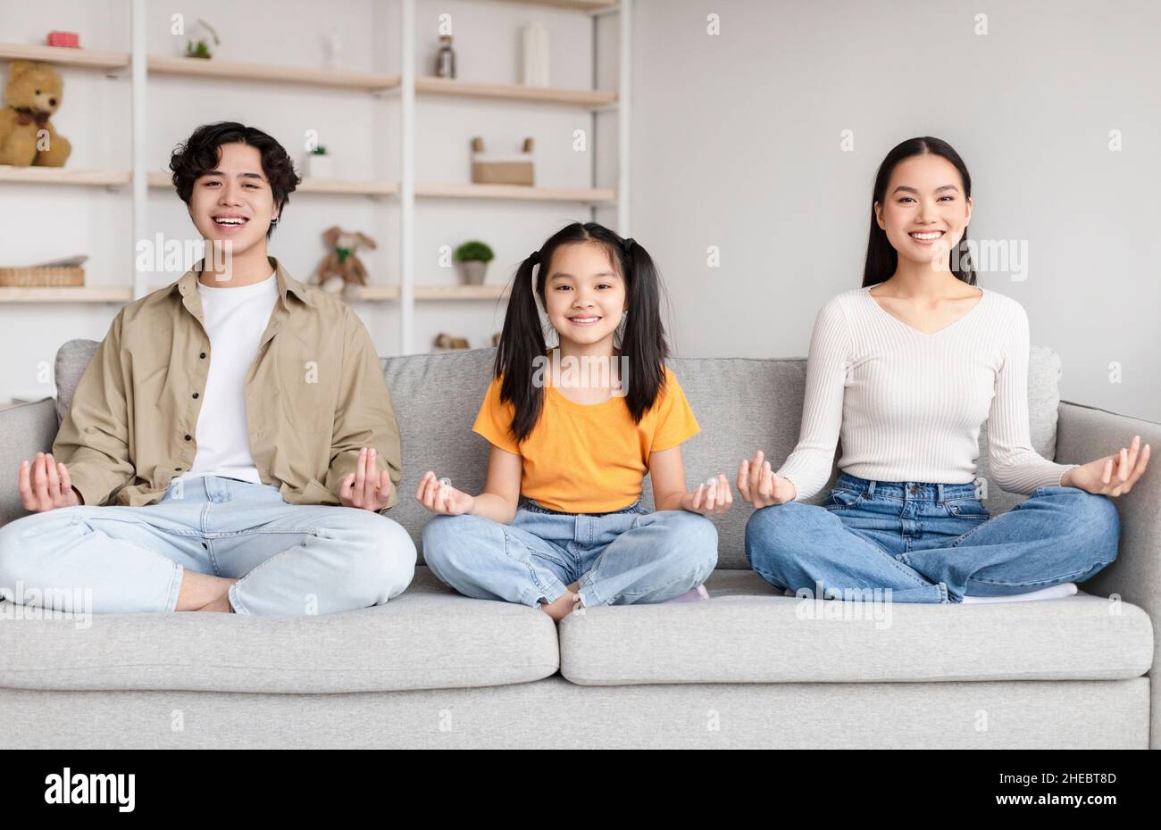 Smiling asian family sits in lotus position, practice yoga on sofa in minimalist living room interior at free time Stock Photo