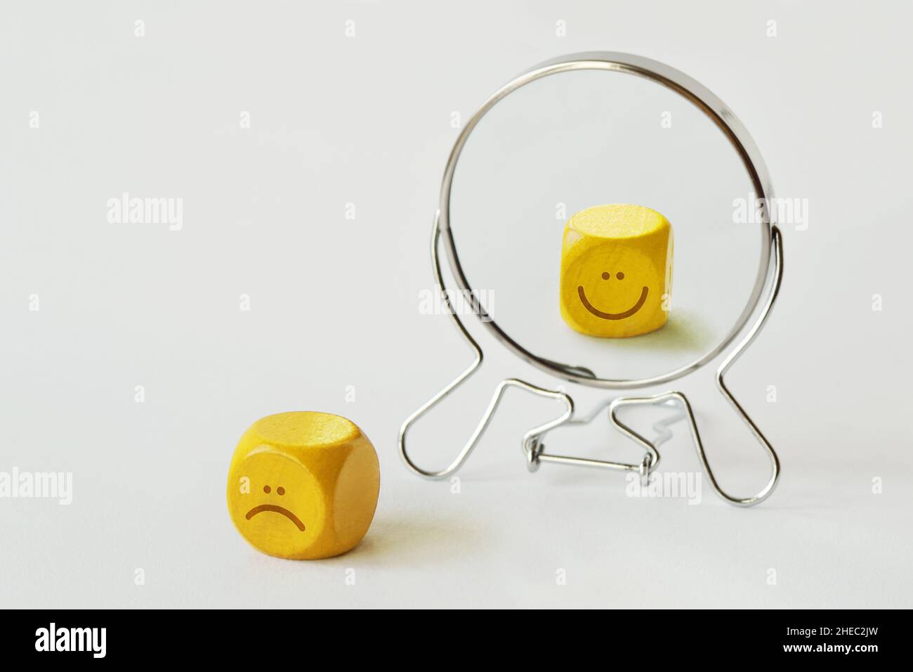 Yellow dice with sad expression looking in the mirror and seeing himself smiling - Concept of person who hides his pain behind his smile Stock Photo