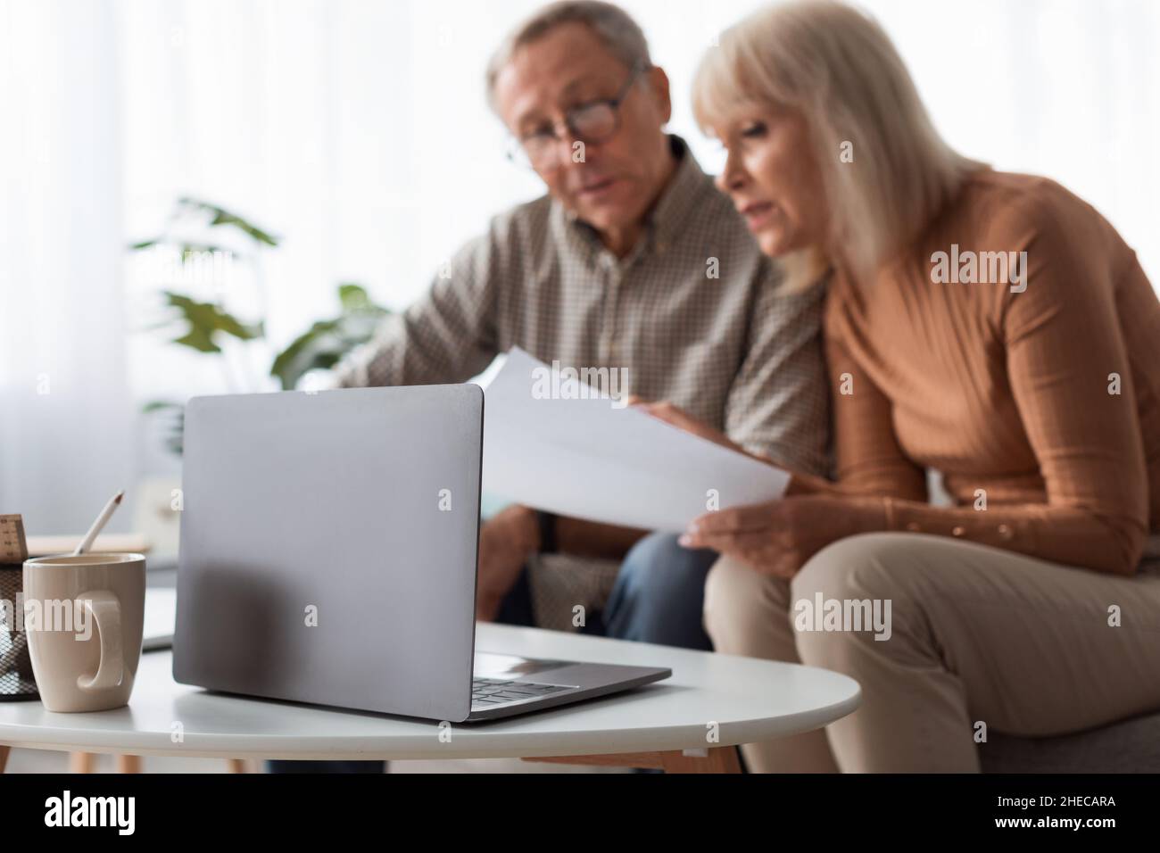 Senior Couple Reading Papers Using Laptop Sitting On Couch Indoors Stock Photo