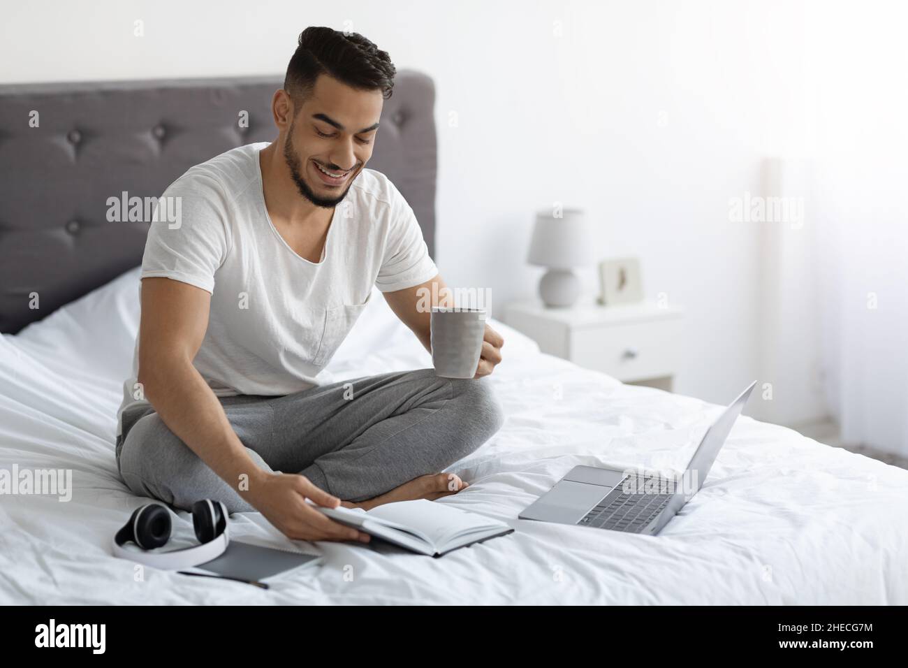 Remote Work. Arab Freelancer Man Working With Laptop And Notepad In Bed Stock Photo