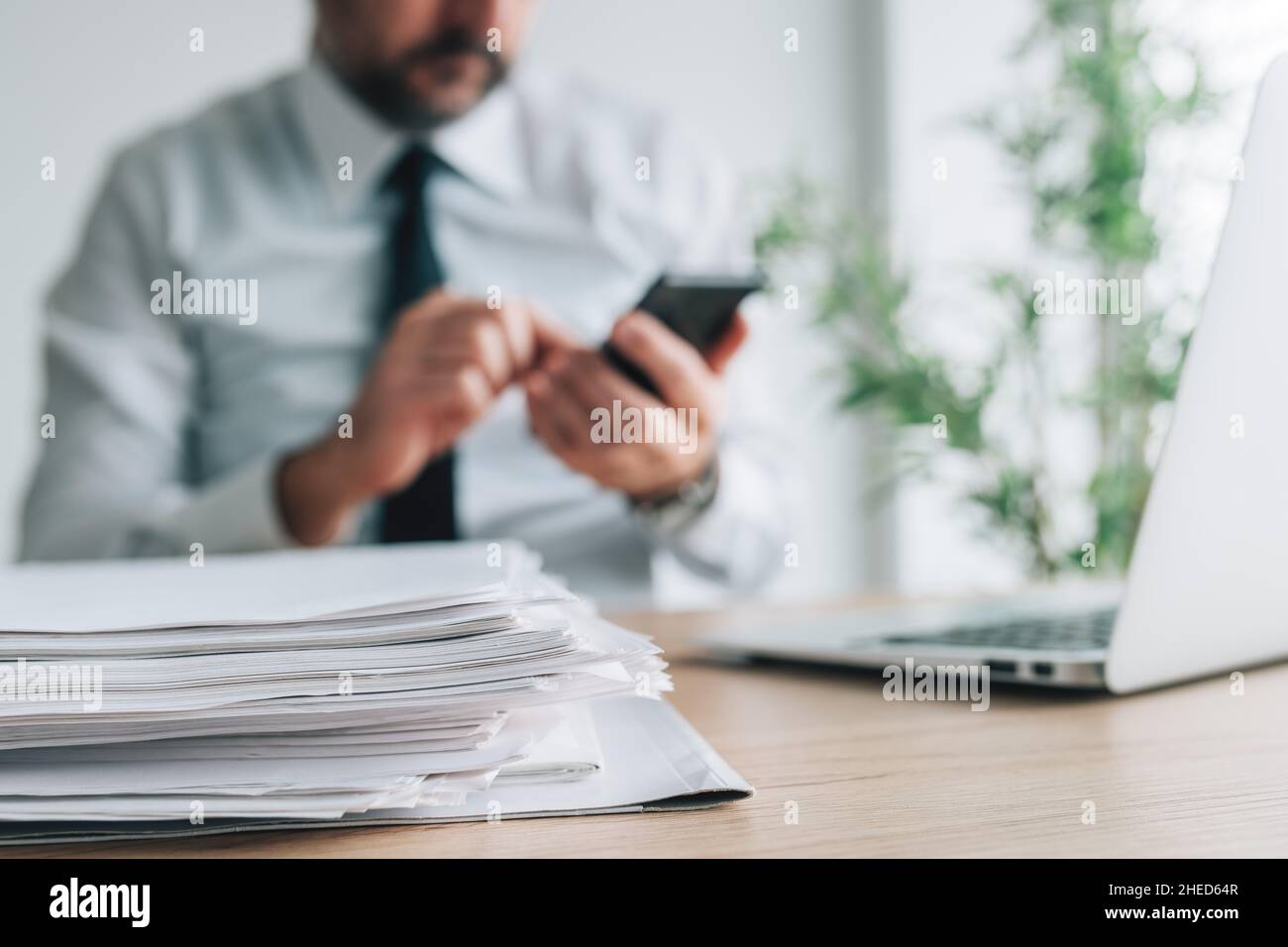 Boreout syndrome, bored businessman using smartphone while paperwork is piling on office desk, selective focus Stock Photo