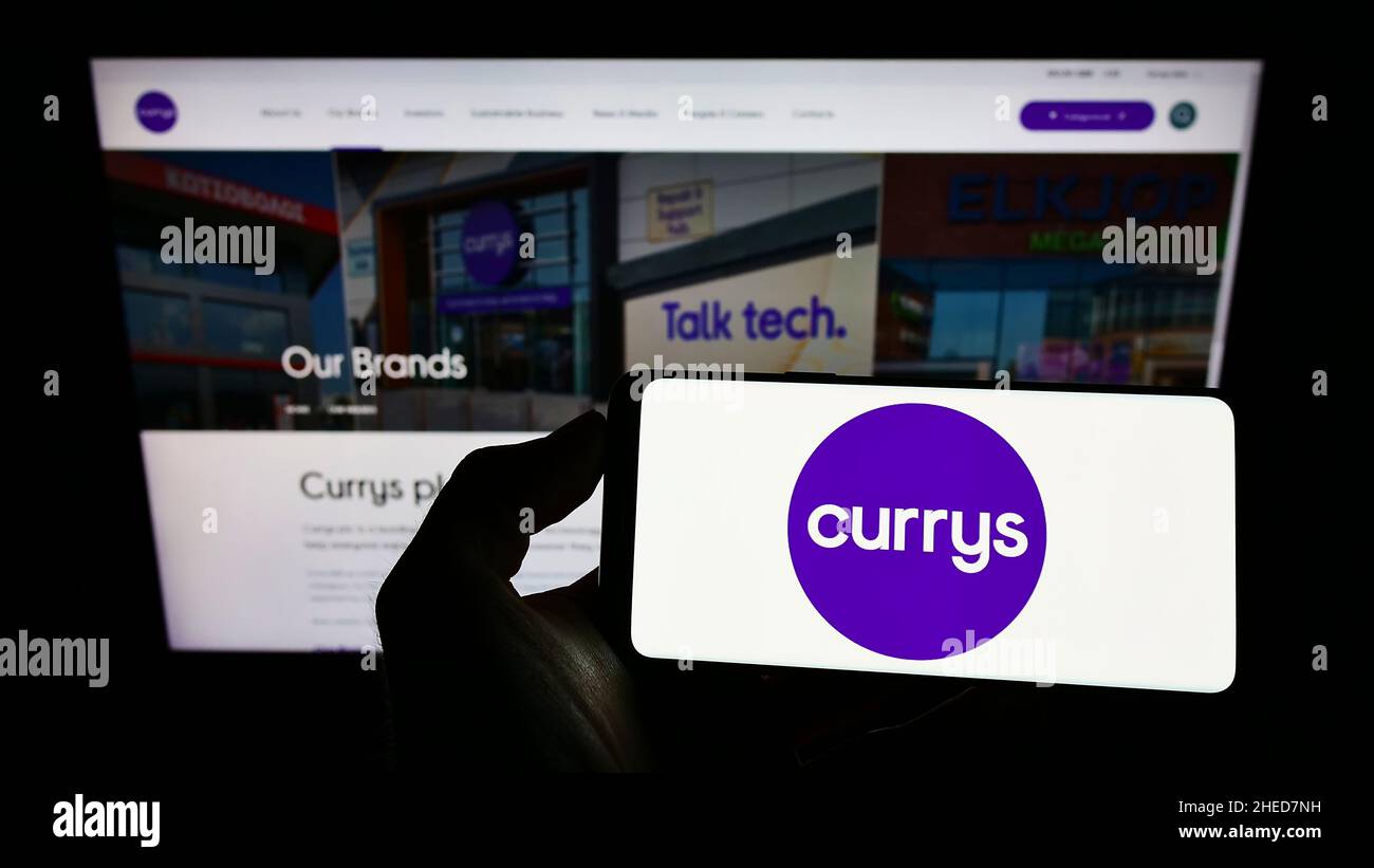 Person holding smartphone with logo of British electronics retail company Currys plc on screen in front of website. Focus on phone display. Stock Photo