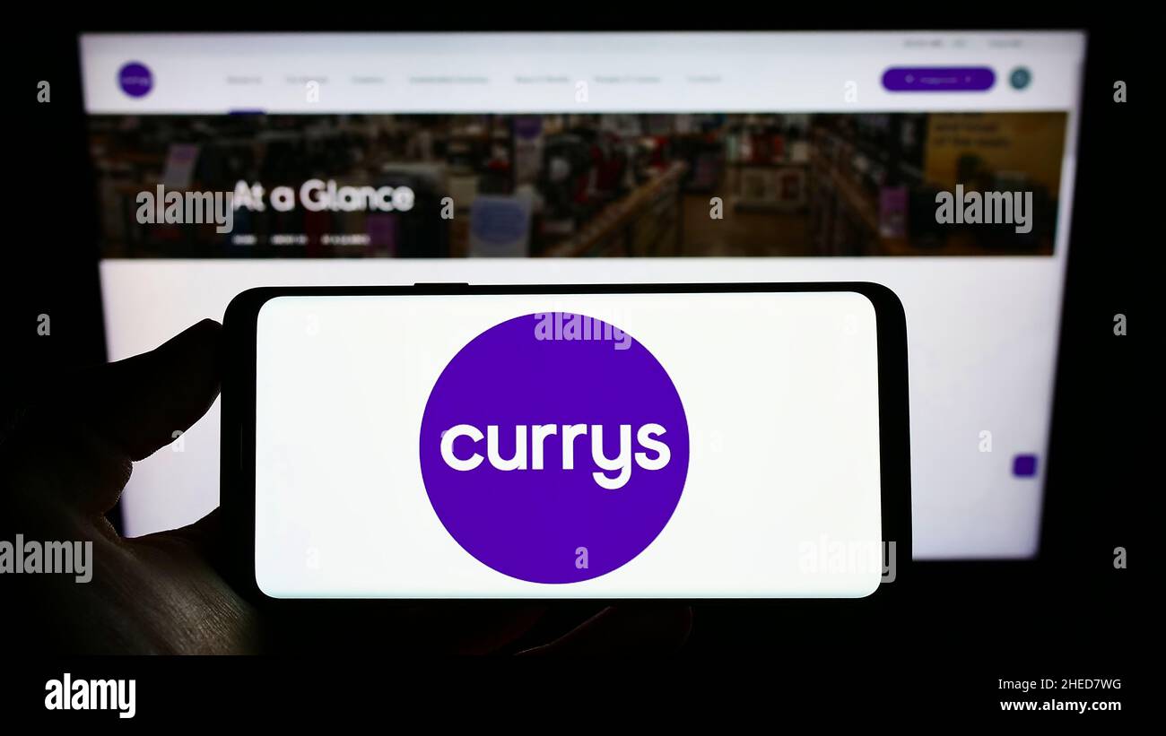 Person holding cellphone with logo of British electronics retail company Currys plc on screen in front of webpage. Focus on phone display. Stock Photo