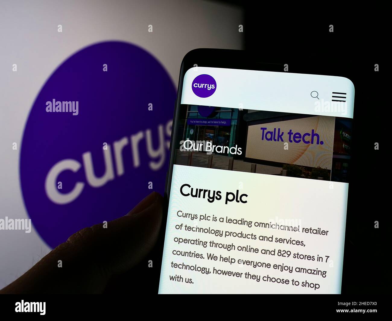 Person holding cellphone with webpage of electronics retail company Currys plc on screen in front of logo. Focus on center of phone display. Stock Photo