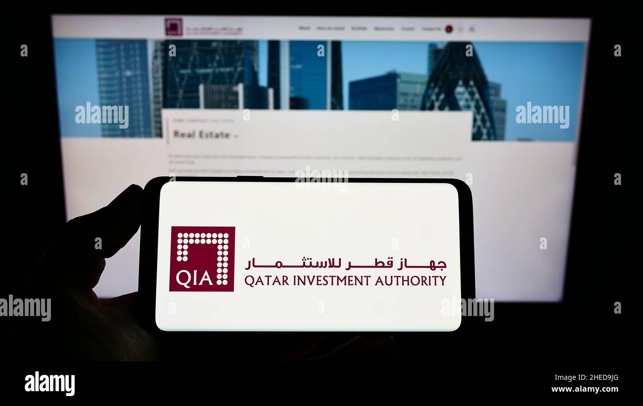 Person holding mobile phone with logo of wealth fund Qatar Investment Authority (QIA) on screen in front of web page. Focus on phone display. Stock Photo