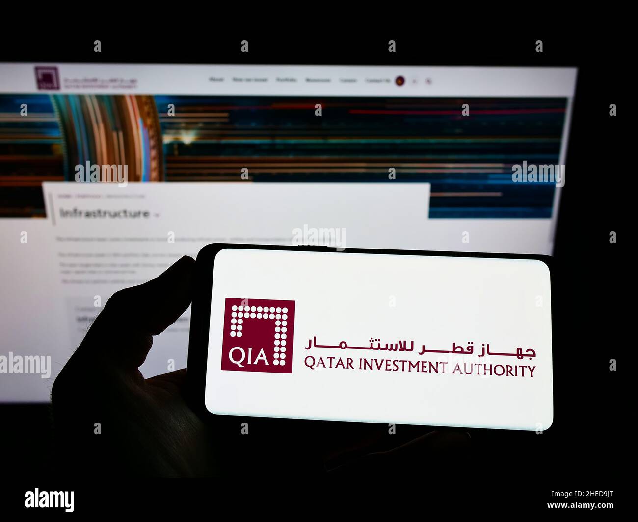 Person holding cellphone with logo of wealth fund Qatar Investment Authority (QIA) on screen in front of business webpage. Focus on phone display. Stock Photo