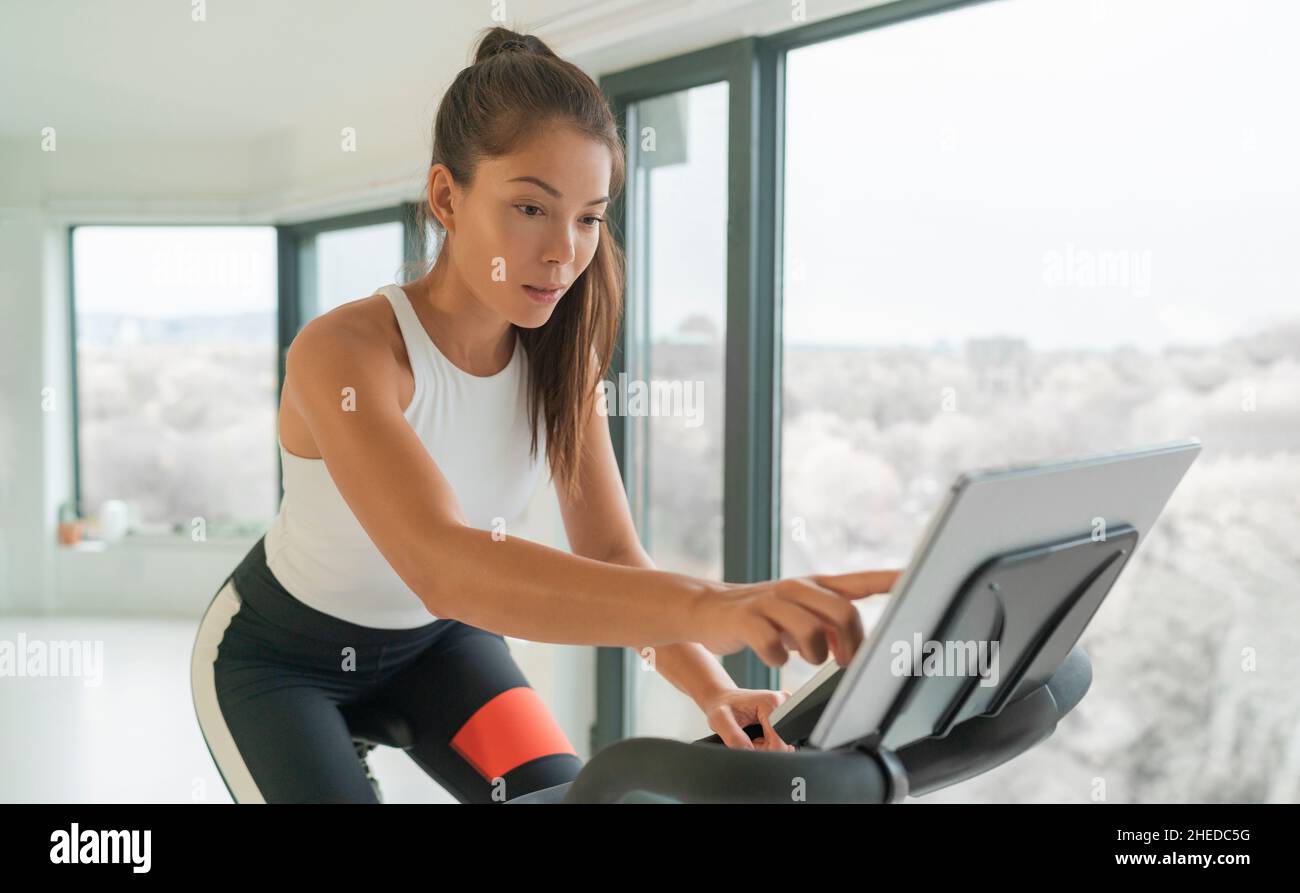 Home cycle workout Asian fit athlete woman training on stationary bike doing online cycling exercise class biking during livestream on screen Stock Photo