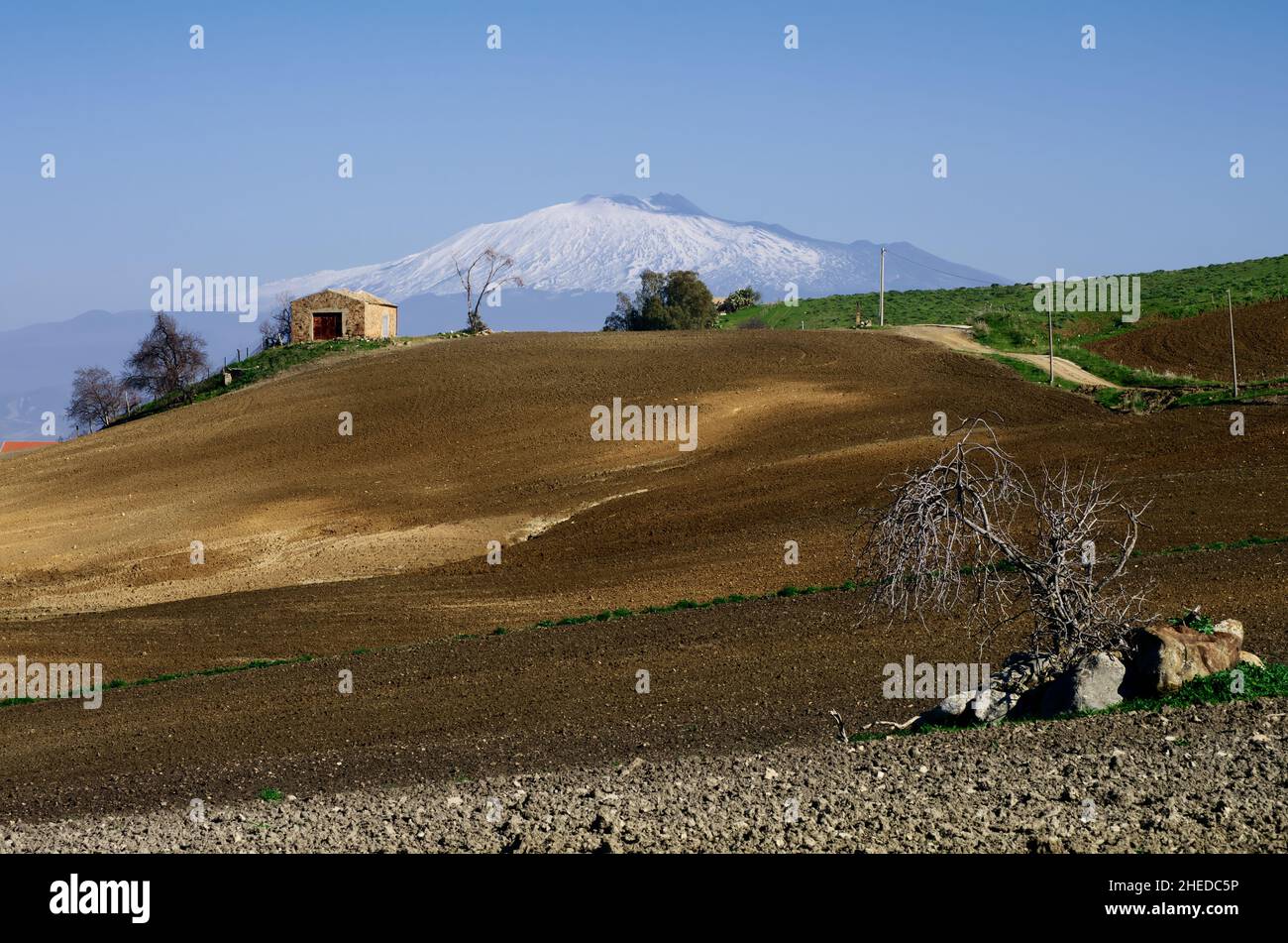 plowed field with a rural house on a hill in the sicilian landscape on the background Etna Mount snow covered Stock Photo