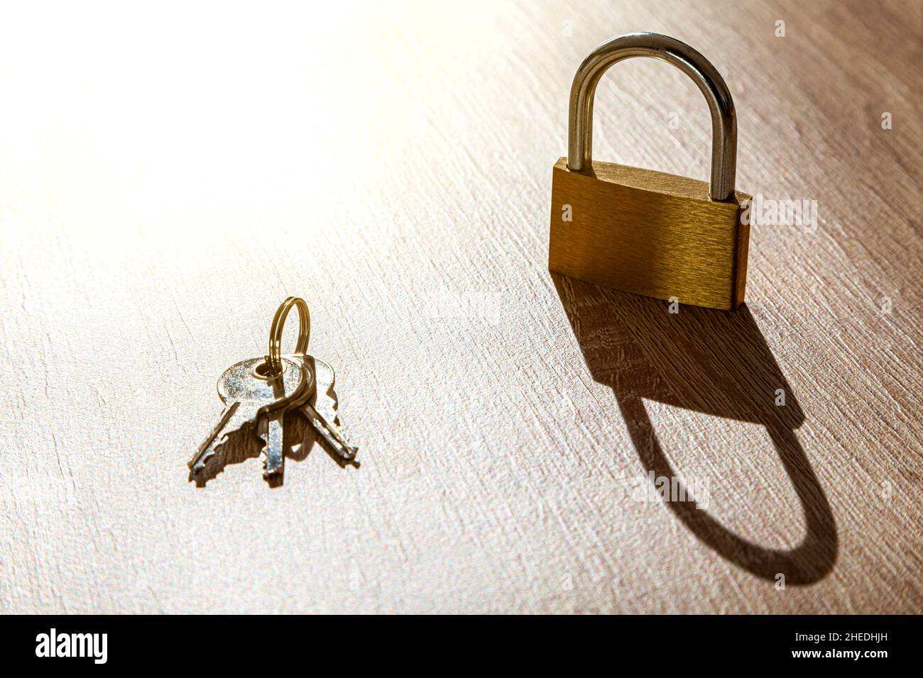 On a wooden table there is a padlock planted and next to it a ring with three keys. The light comes intense from above and casts a hard, elongated sha Stock Photo