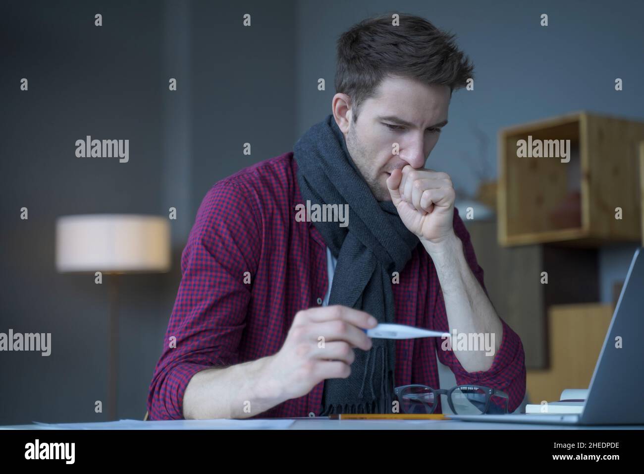 Unhealthy looking male entrepreneur in scarf around his neck coughing looks at digital thermometer Stock Photo