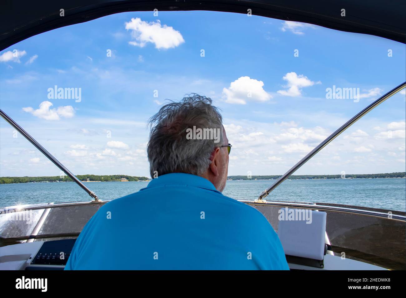 Back close-up view of grey haired man in sunglasses driving a boat over the lake with houses and shoreline on horizon under blue cloudy sky Stock Photo