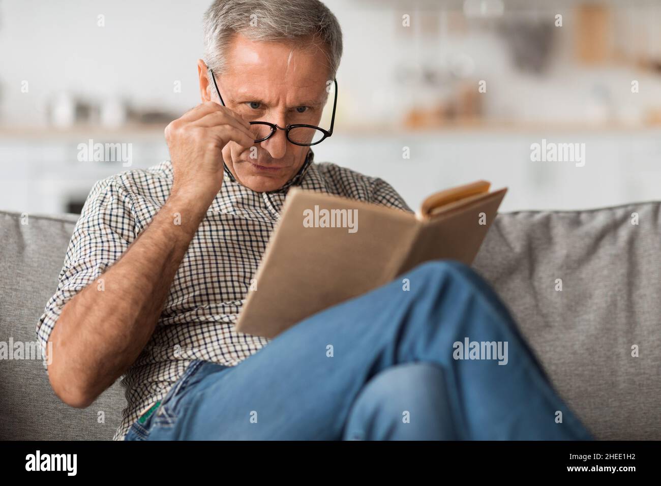 Senior Man With Poor Eyesight Reading Book Above Eyeglasses Indoor Stock Photo