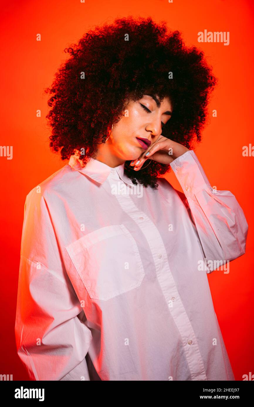 Self assured young Hispanic female model in white shirt touching face with eyes closed in studio with bright red neon illumination Stock Photo