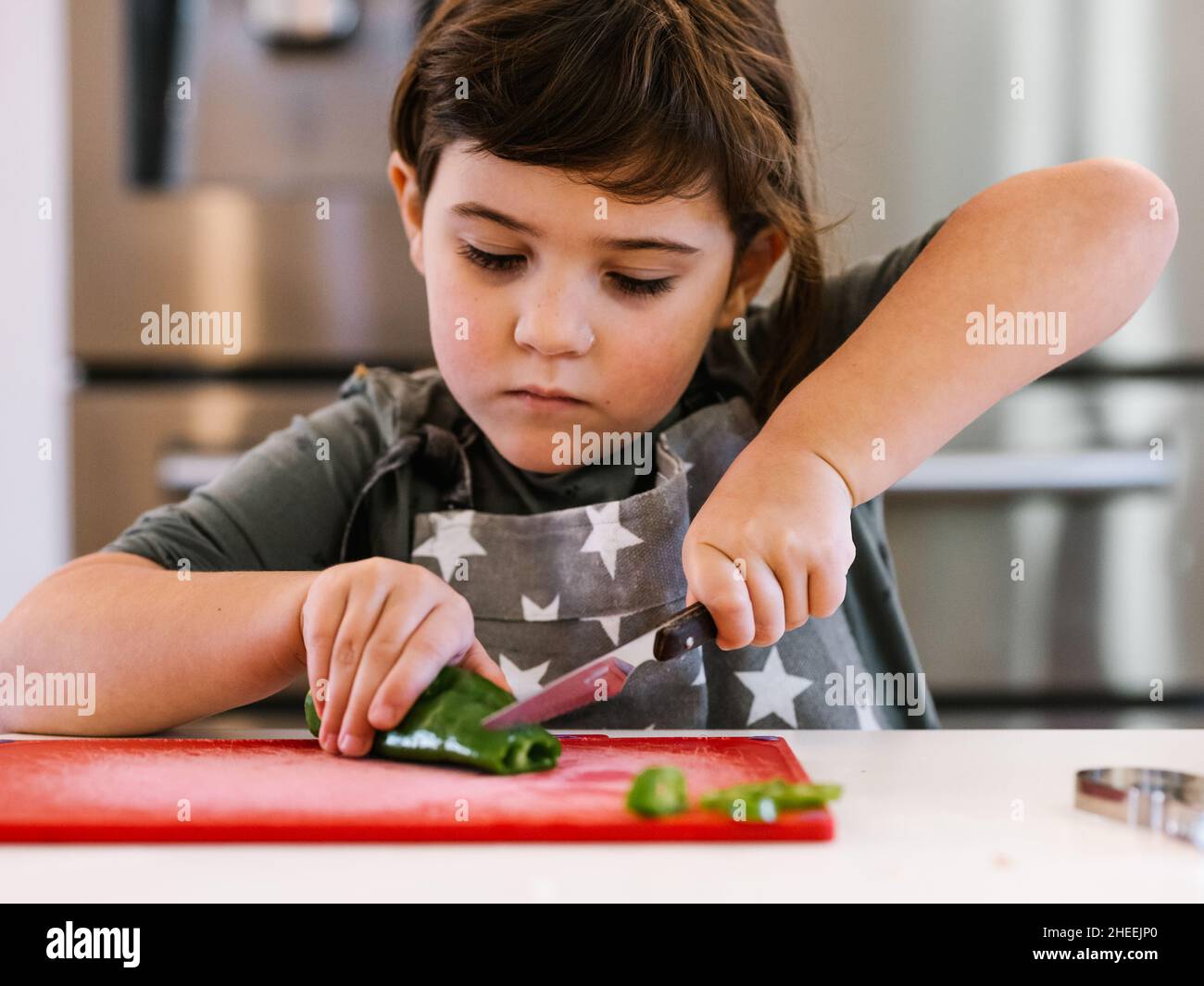 Focused girl with brown hair and in apron slicing fresh green pepper on