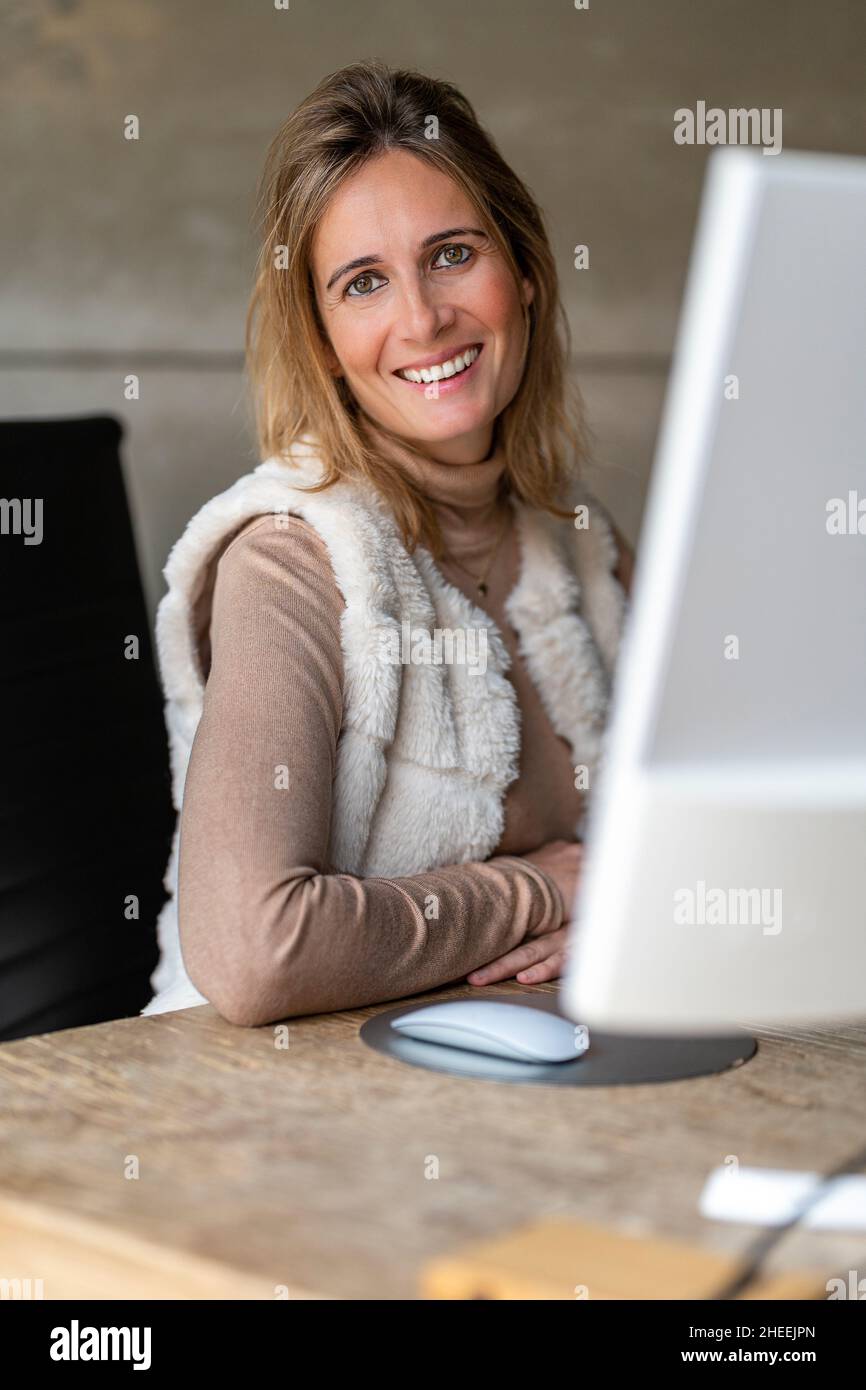 Cheerful female worker typing on laptop while looking at camera in light modern workspace Stock Photo