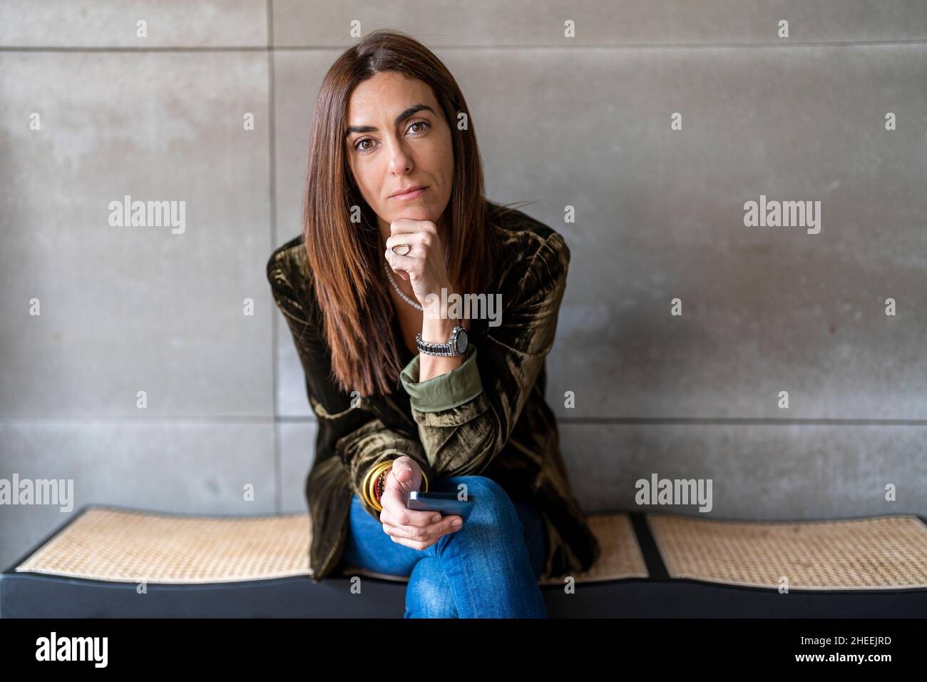Focused female entrepreneur with cellphone while sitting on bench at gray wall in light modern office during workday Stock Photo