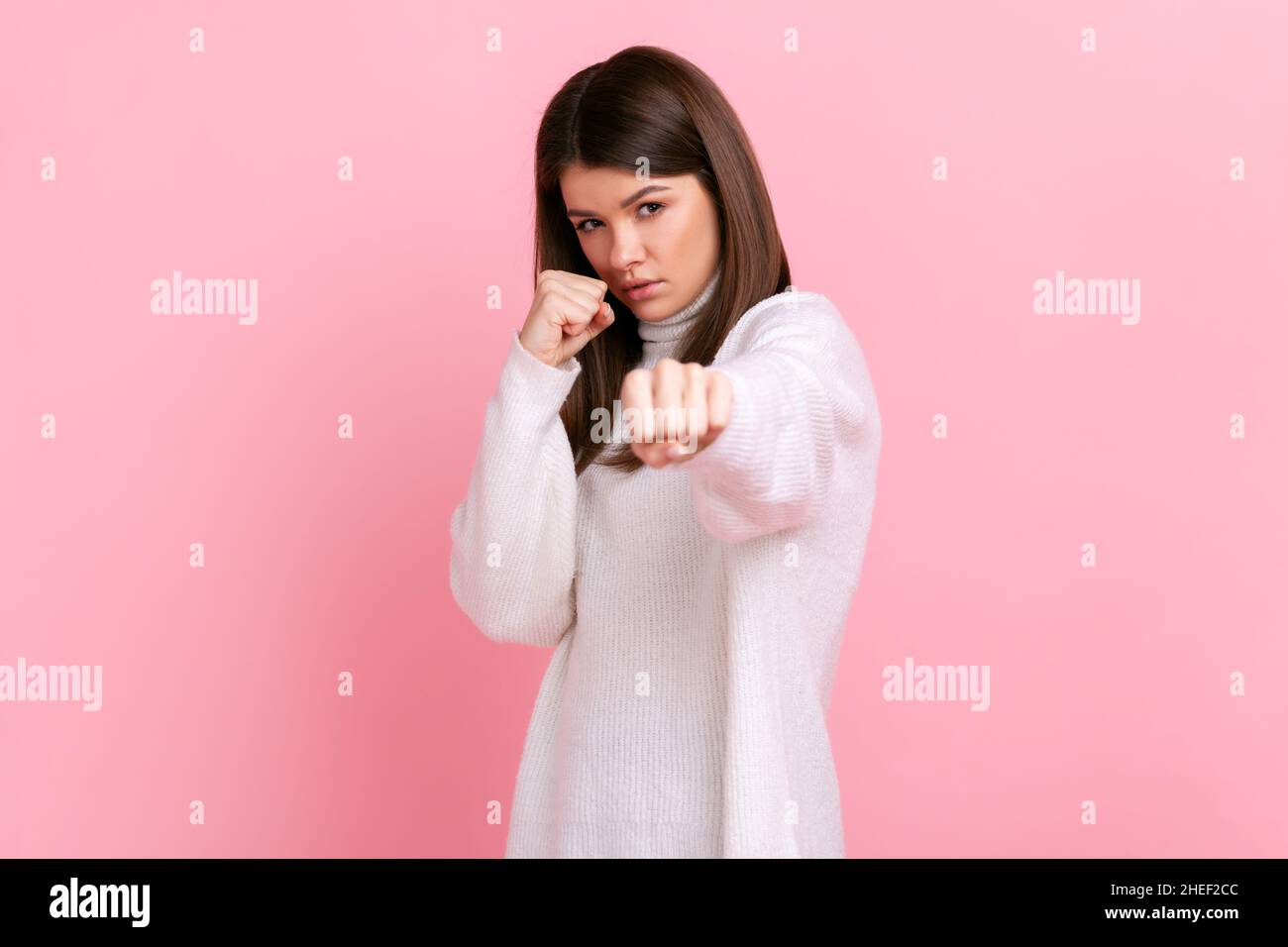 Aggressive angry woman with dark hair being ready to fight, boxing to camera with clenched fists, wearing white casual style sweater. Indoor studio shot isolated on pink background. Stock Photo