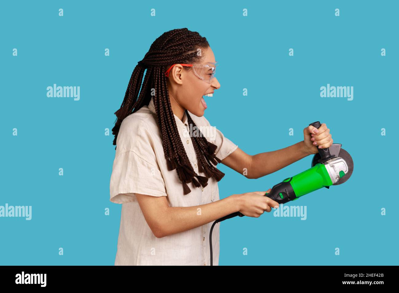 Side view of enthusiastic woman with dreadlocks working with grinder saw, yelling with excitement, wearing white shirt and protective glasses. Indoor studio shot isolated on blue background. Stock Photo