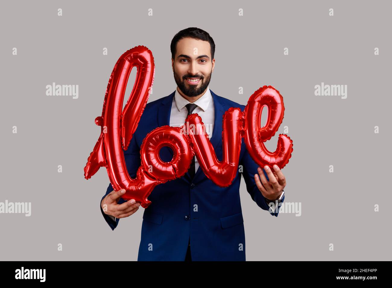 Bearded businessman holding love word of foil balloons, expressing positive emotions and his feelings, wearing official style suit. Indoor studio shot isolated on gray background. Stock Photo