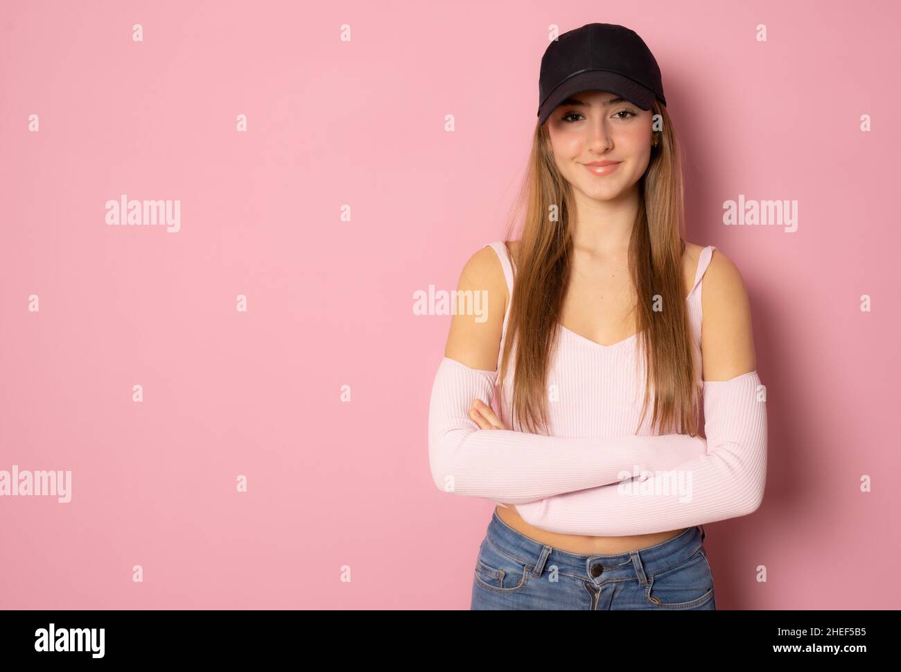 Beautiful girl wearing casual clothing and black cap posing with arms folded isolated over pink background. Stock Photo