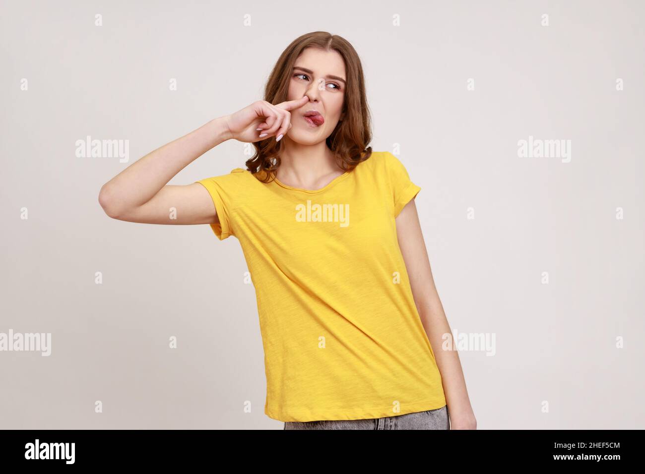 Funny comical teenager girl with brown hair picking finger to nose and showing tongue, looking at away, having fun, fooling around, bad manners. Indoor studio shot isolated on gray background. Stock Photo