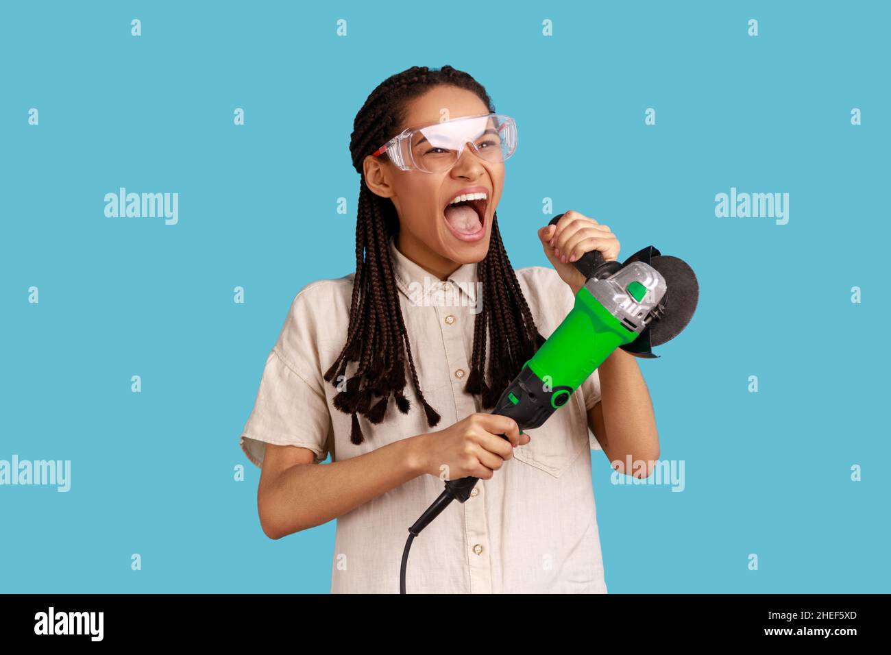 Portrait of enthusiastic woman with dreadlocks working with grinder saw, yelling with excitement, wearing white shirt and protective glasses. Indoor studio shot isolated on blue background. Stock Photo