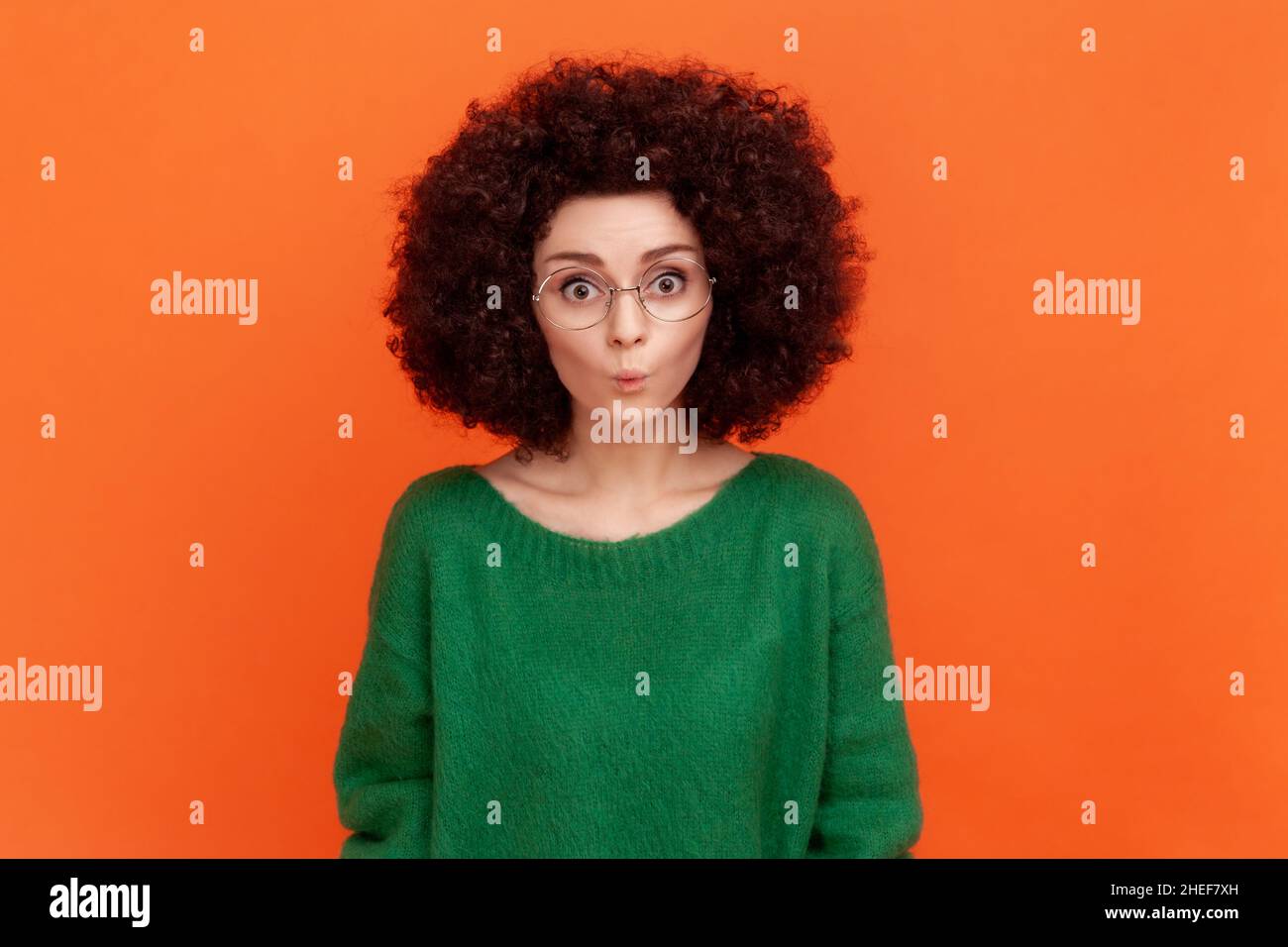 Portrait of funny woman with Afro hairstyle wearing green casual style sweater and eyeglasses, standing with pout lips, looking at camera. Indoor studio shot isolated on orange background. Stock Photo