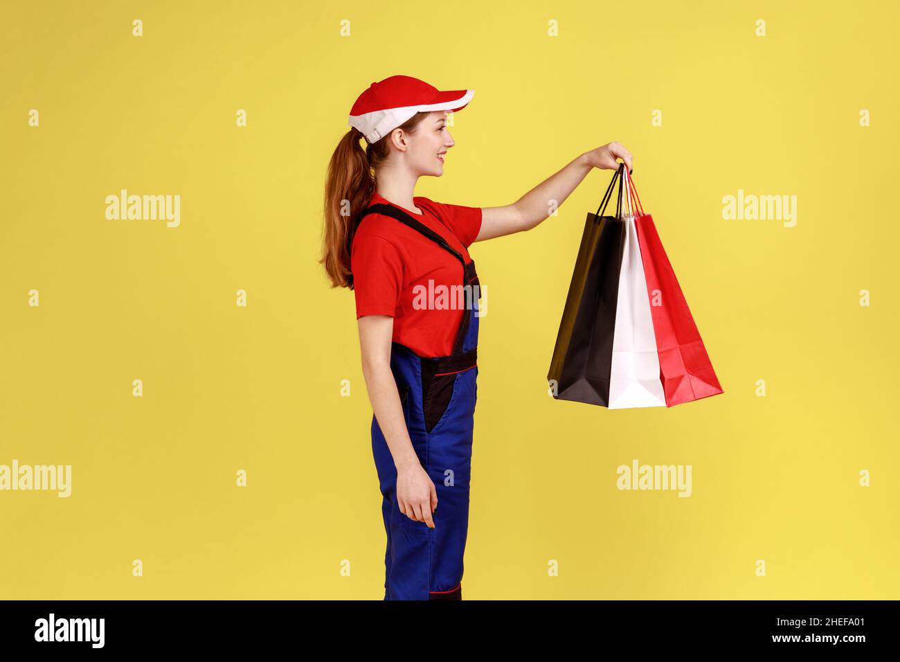 Side view portrait of optimistic courier woman standing with shopping bags in hands, expressing happiness, wearing overalls and red cap. Indoor studio shot isolated on yellow background. Stock Photo
