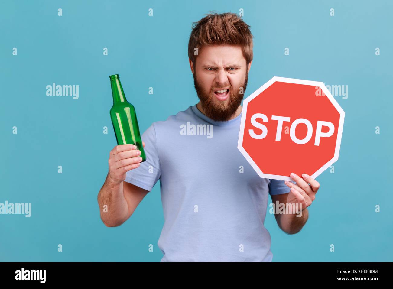 Portrait of bearded man showing alcoholic beverage beer bottle and stop sign, warning and worrying, looking at camera with angry expression. Indoor studio shot isolated on blue background. Stock Photo