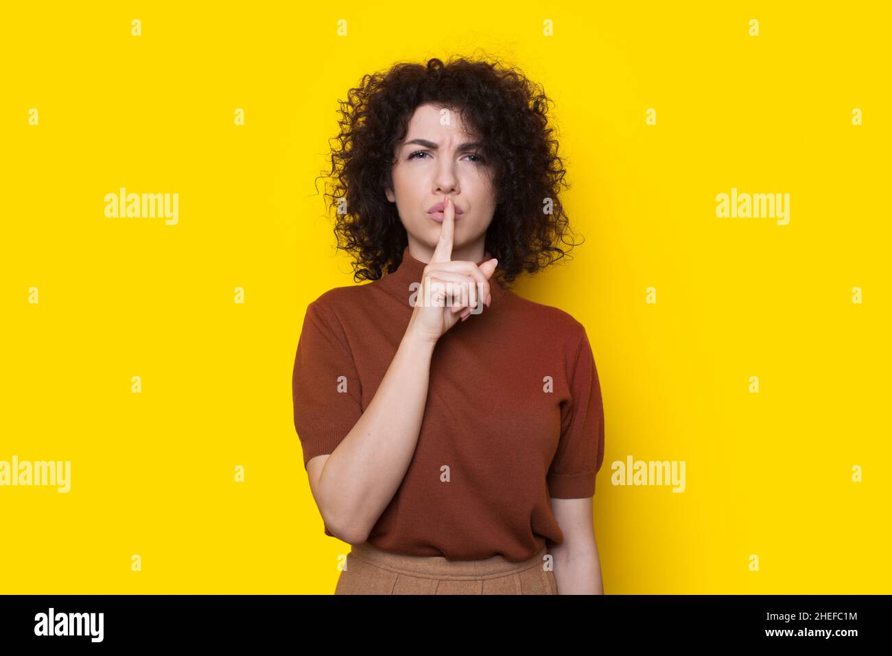 Portrait of serious woman showing silence gesture with finger over lips isolated over yellow background. Shh sign. Curly hair. Stock Photo