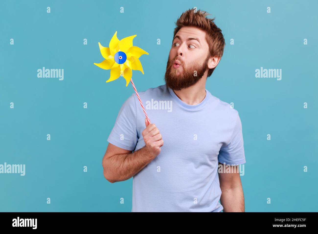 Portrait of funny positive childish handsome bearded man wearing T-shirt playing blowing yellow windmill, having fun with paper toy. Indoor studio shot isolated on blue background. Stock Photo