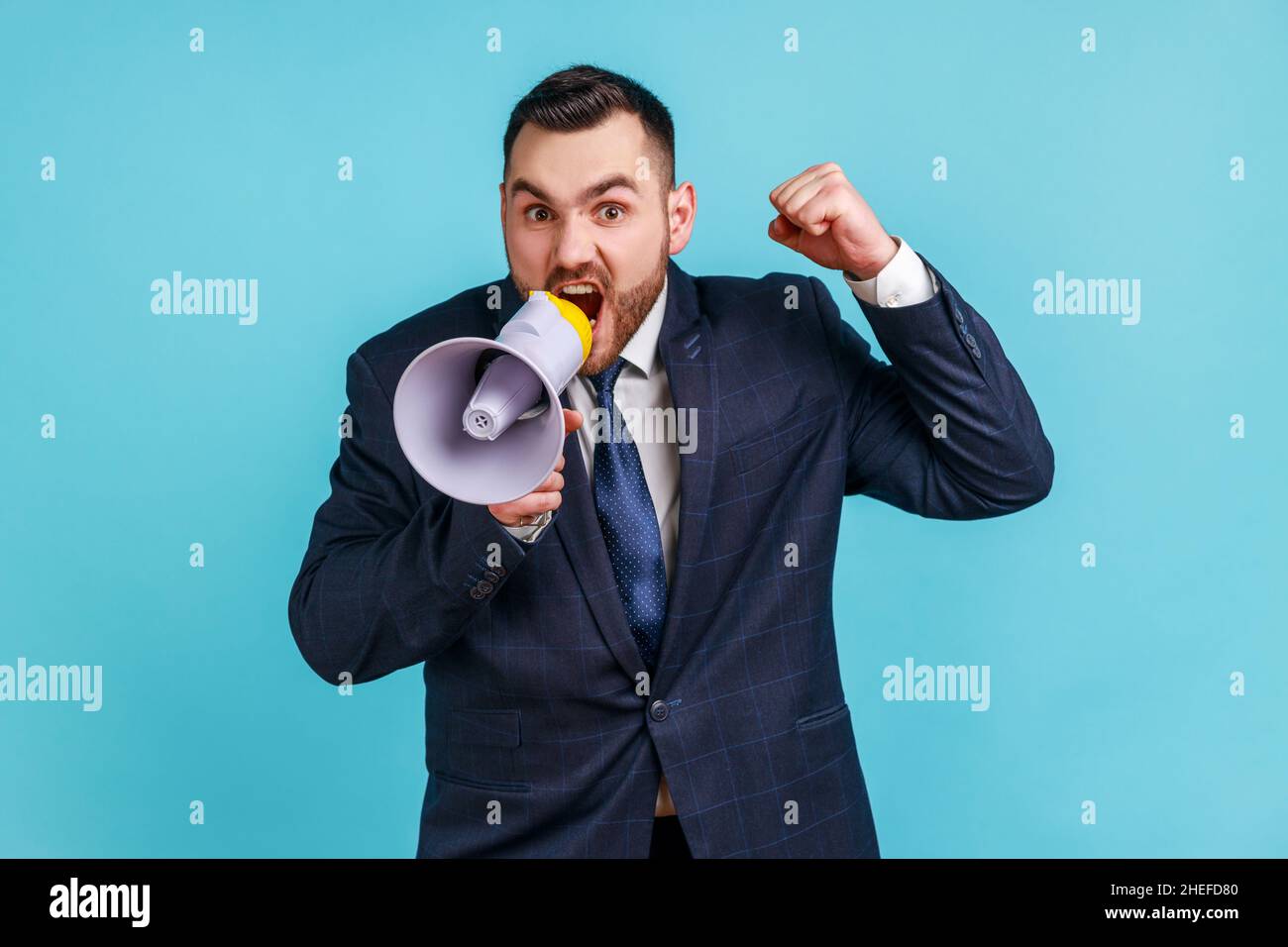 Angry serious businessman wearing dark official style suit loudly speaking screaming holding megaphone, announcing important message. Indoor studio shot isolated on blue background. Stock Photo