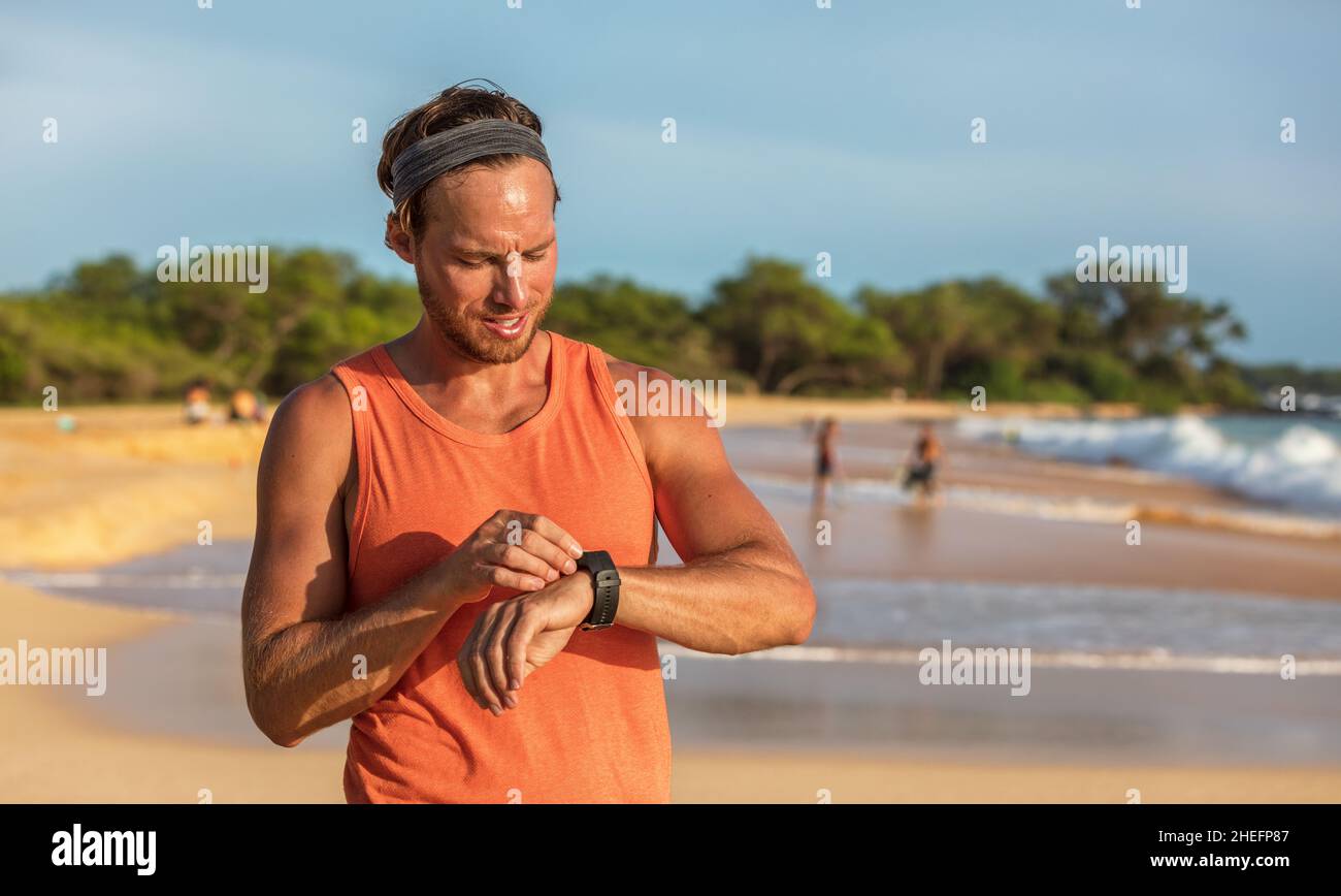 Running man athlete using smartwatch fitness app on wearable tech device smart sports watch training on beach. Runner working out sweating on summer Stock Photo
