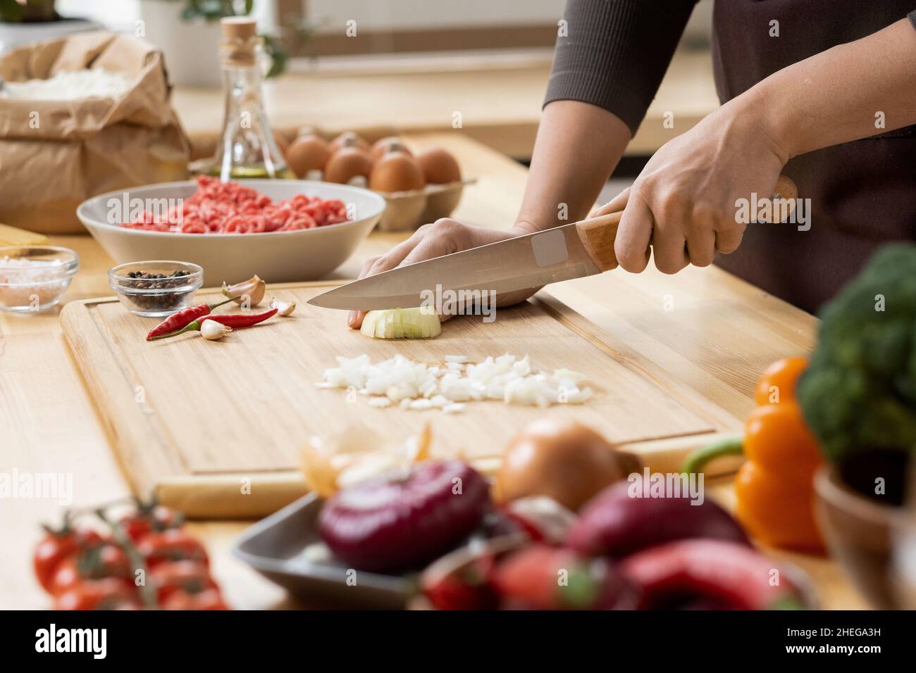 Hands of young female chopping fresh onion on wooden board while preparing italian pasta with vegetables and minced meat Stock Photo