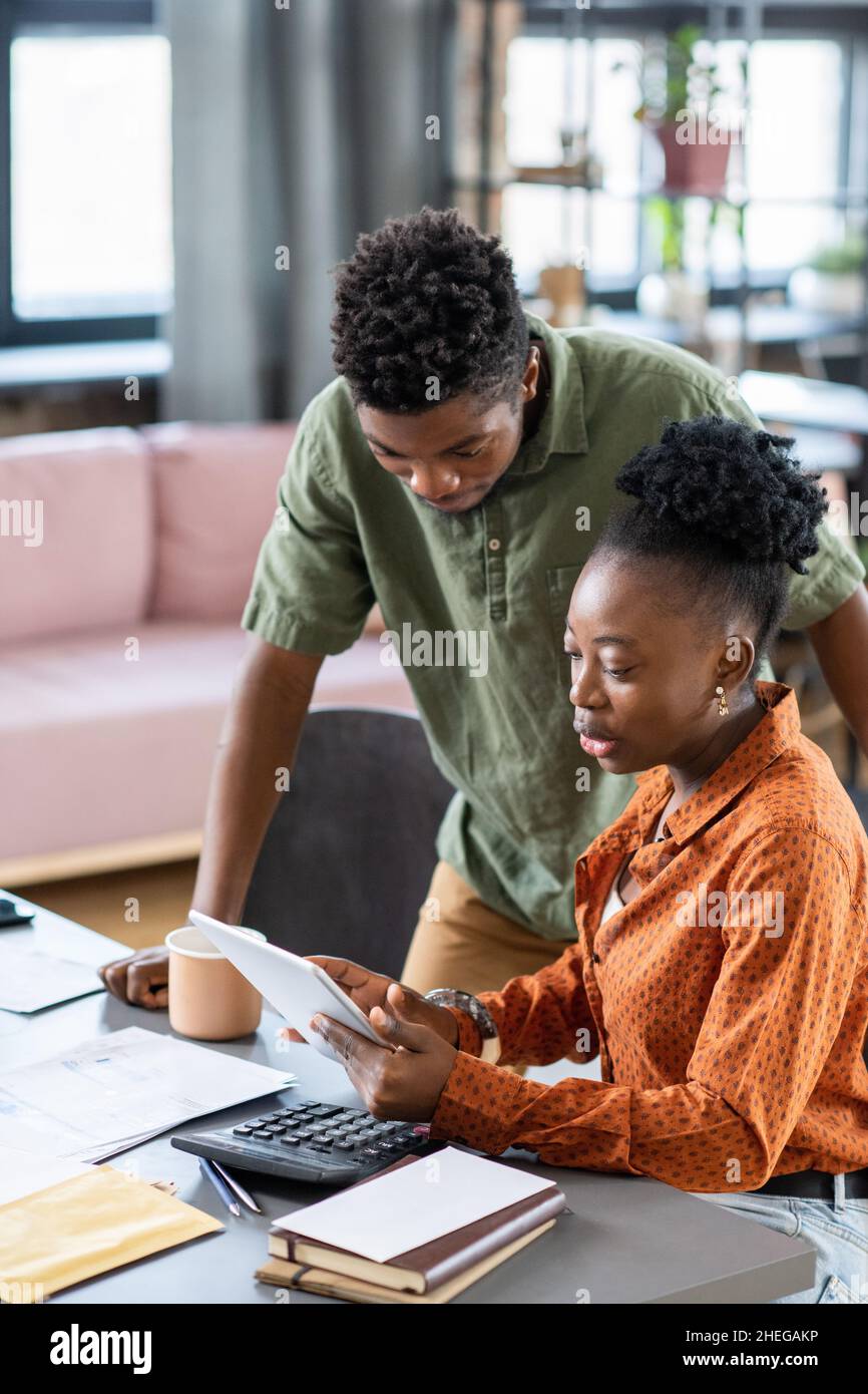 Young African man bending over female colleague with tablet making presentation of online financial data and rate Stock Photo