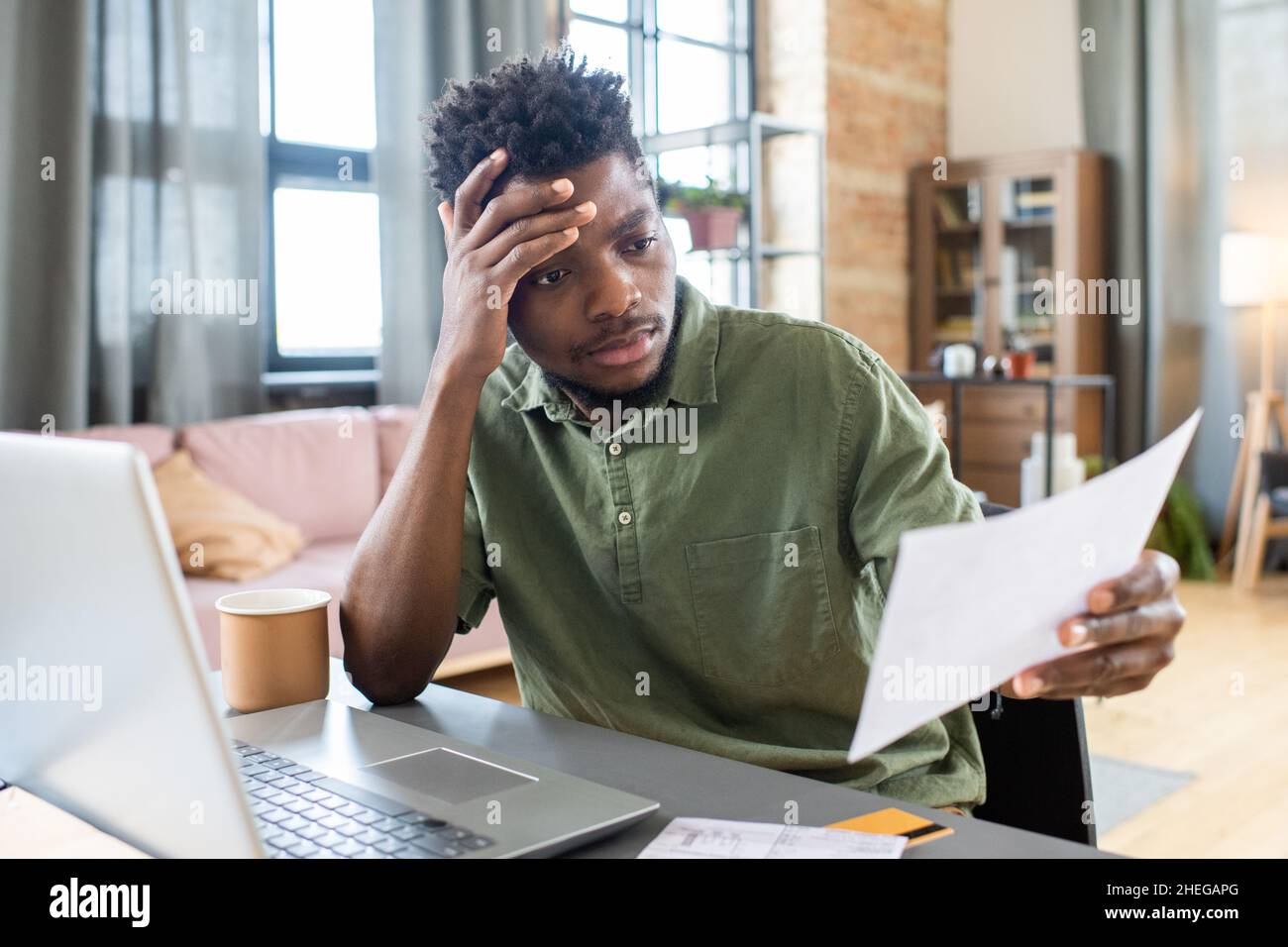 Young stressed African businessman looking through unpaid bill and touching his head while sitting in front of laptop Stock Photo