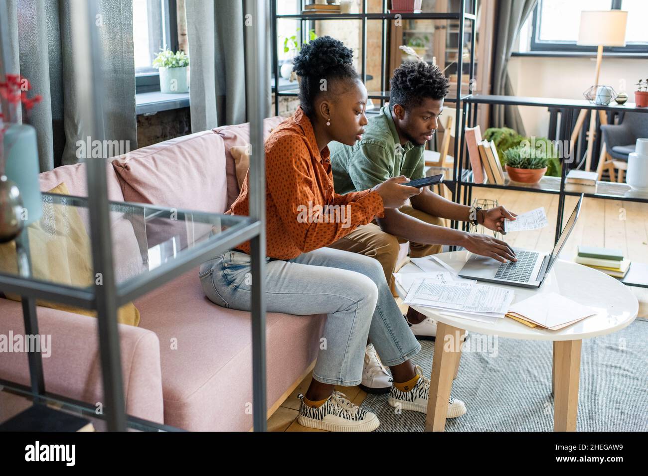 Two young contemporary coworkers working with mobile gadgets and financial data while sitting on couch by table Stock Photo