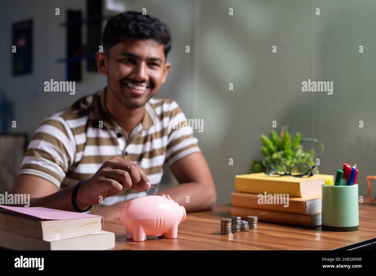Focus on piggy money bank young indian student saving money for future abroad education by placing money on piggy bank while reading at home - concept Stock Photo