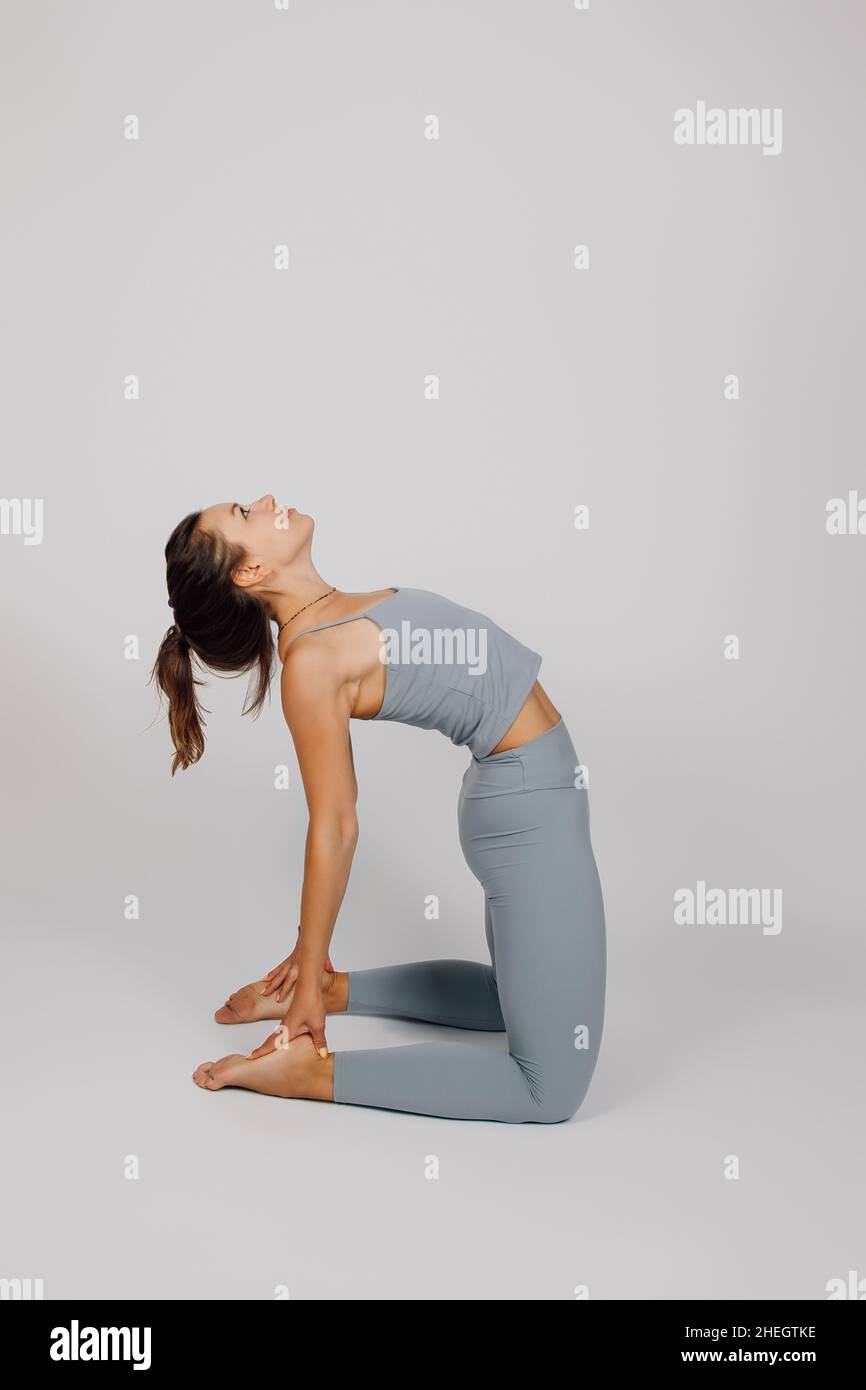 Brunette poses on isolated background. Young woman in gray tracksuit for fitness or yoga does stretching exercise, kneels and holds her heels with her Stock Photo