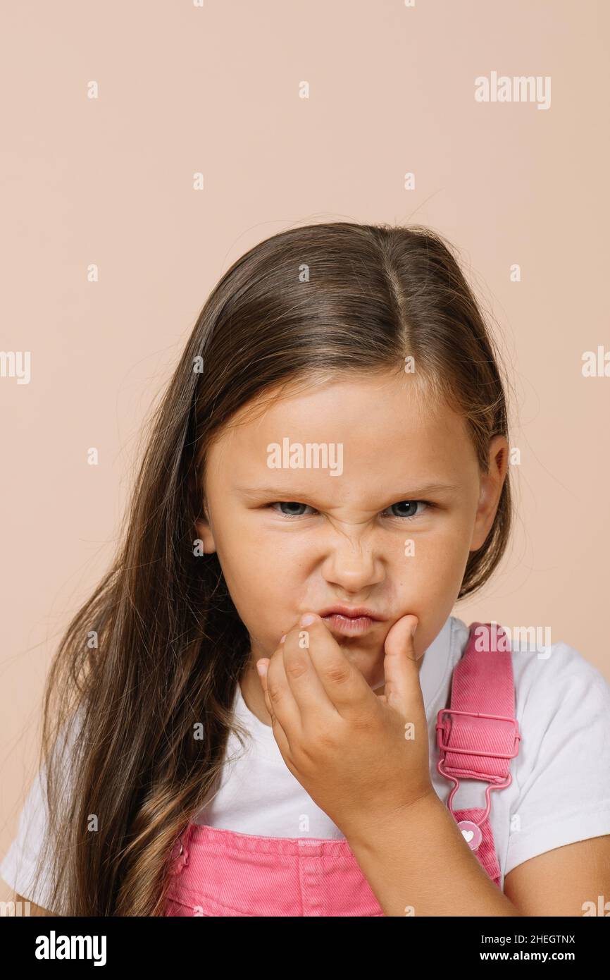 Little girl with dissatisfied grimacing face and displeased eyes touching face with left hand looking at camera wearing bright pink jumpsuit and white Stock Photo