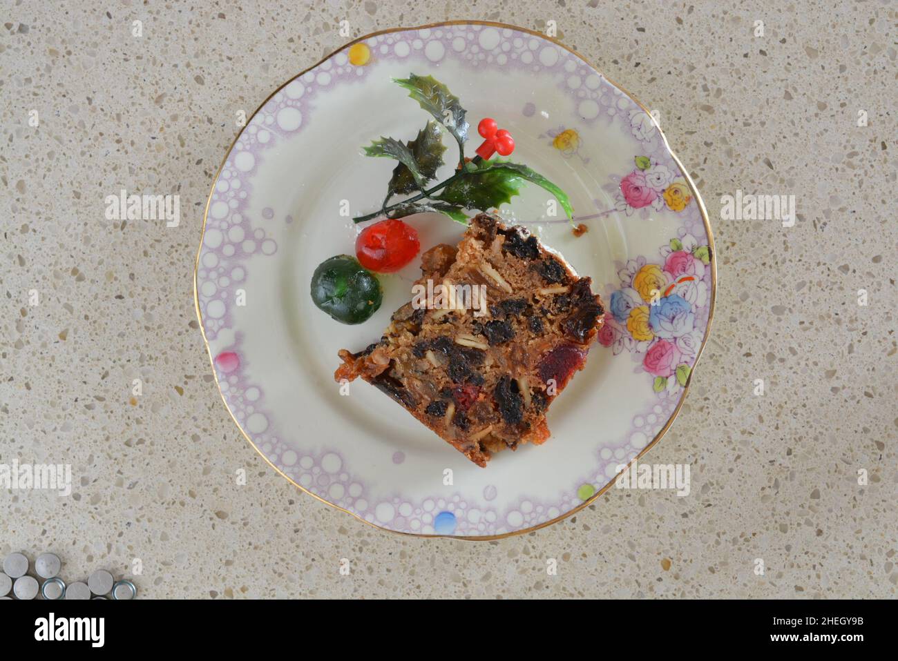 Christmas Cake Slice with cherry and Holly trim on floral cake plate. Decorations in front of cake slice. Stock Photo