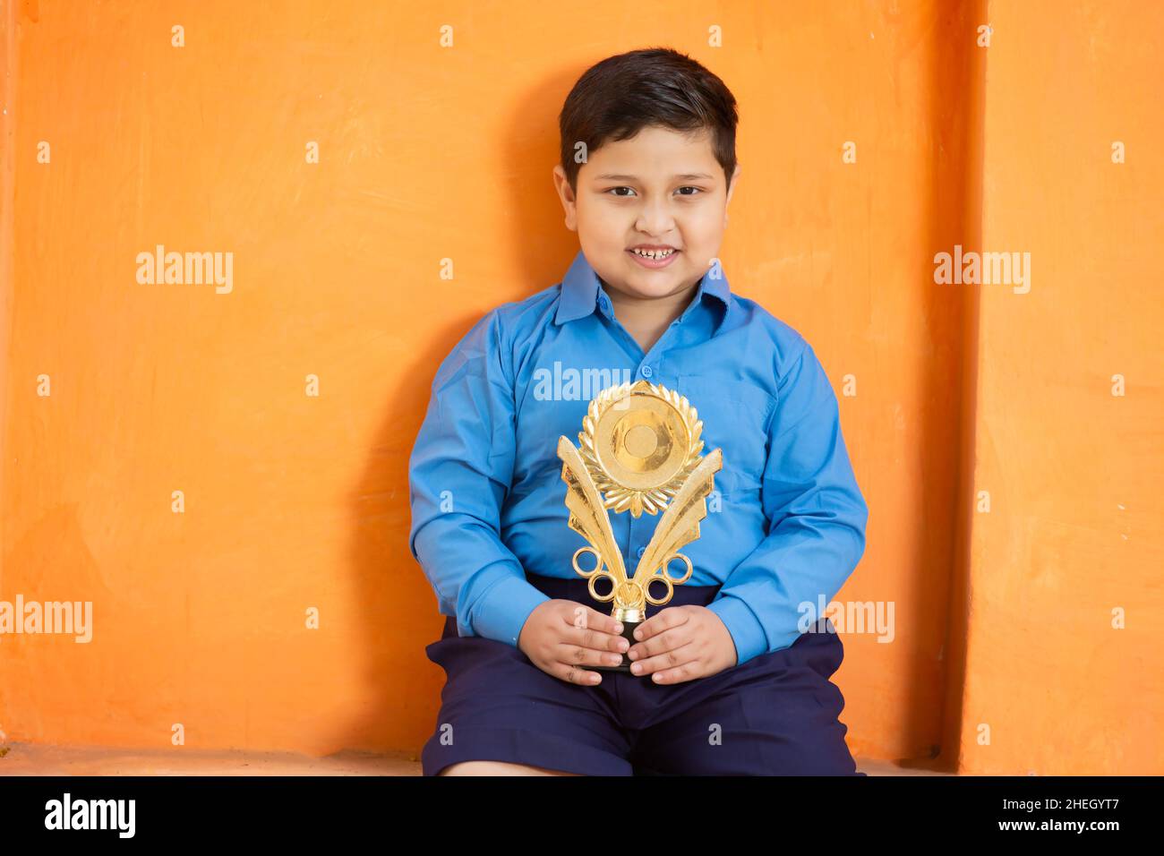 Portrait of happy adorable cute indian boy in school uniform celebrating victory with trophy,Cheerful male kid holding winning prize against orange ba Stock Photo